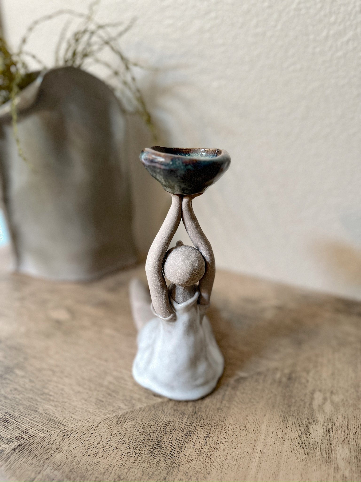 Glazed and Fired ceramic handmade sculpture of a lady holding a small blue/green bowl above her head and sitting on a table. Image from the back.