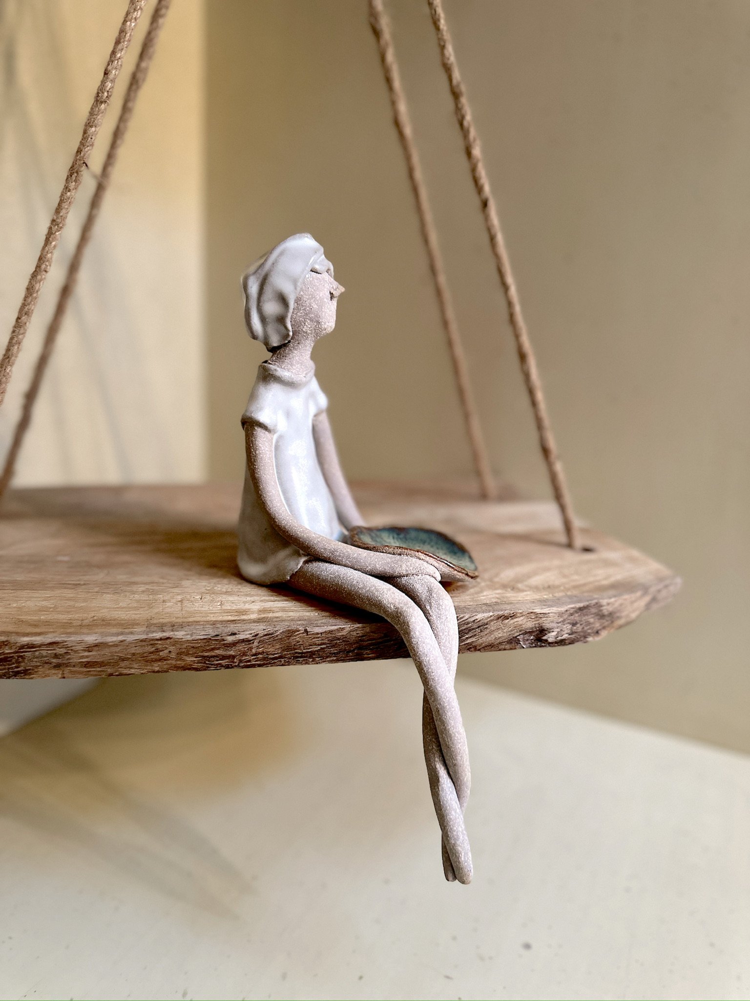 Glazed and Fired ceramic handmade sculpture of a lady holding a small blue/green plate and sitting on the edge of a swing. Image from the right.