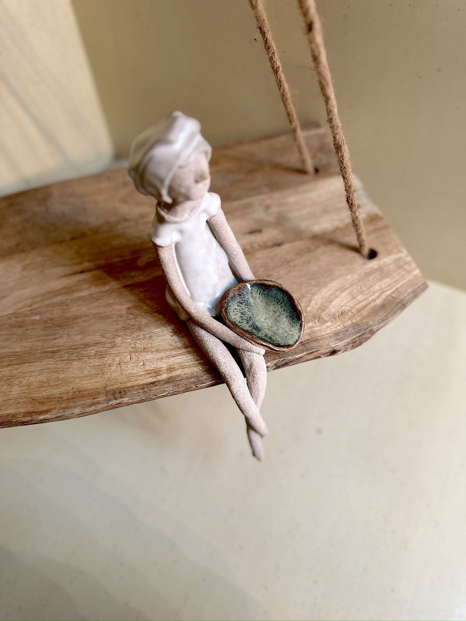 Glazed and Fired ceramic handmade sculpture of a lady holding a small blue/green plate and sitting on the edge of a swing. Image from the top.