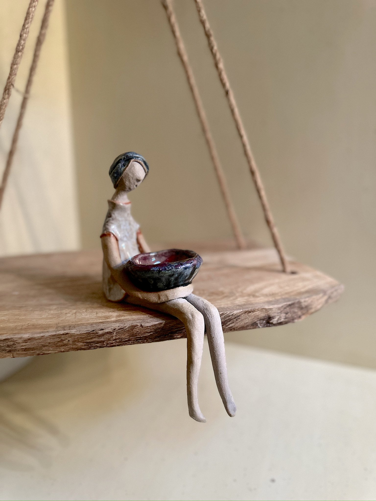 Glazed and Fired ceramic handmade sculpture of a lady holding a red and blue bowl and sitting on the edge of a swing. Image from the right.