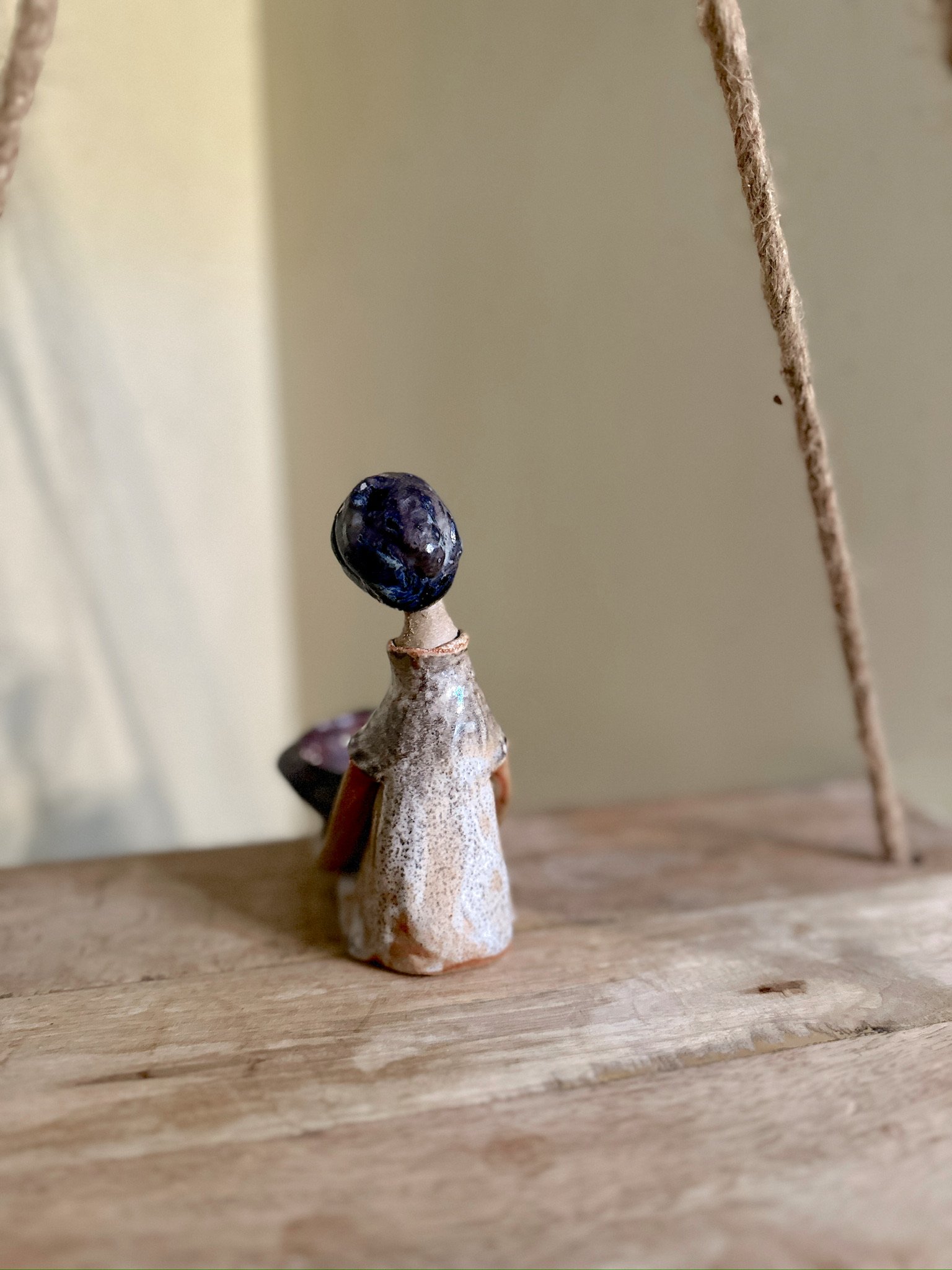 Glazed and Fired ceramic handmade sculpture of a lady holding a red and blue bowl and sitting on the edge of a swing. Image from the back.
