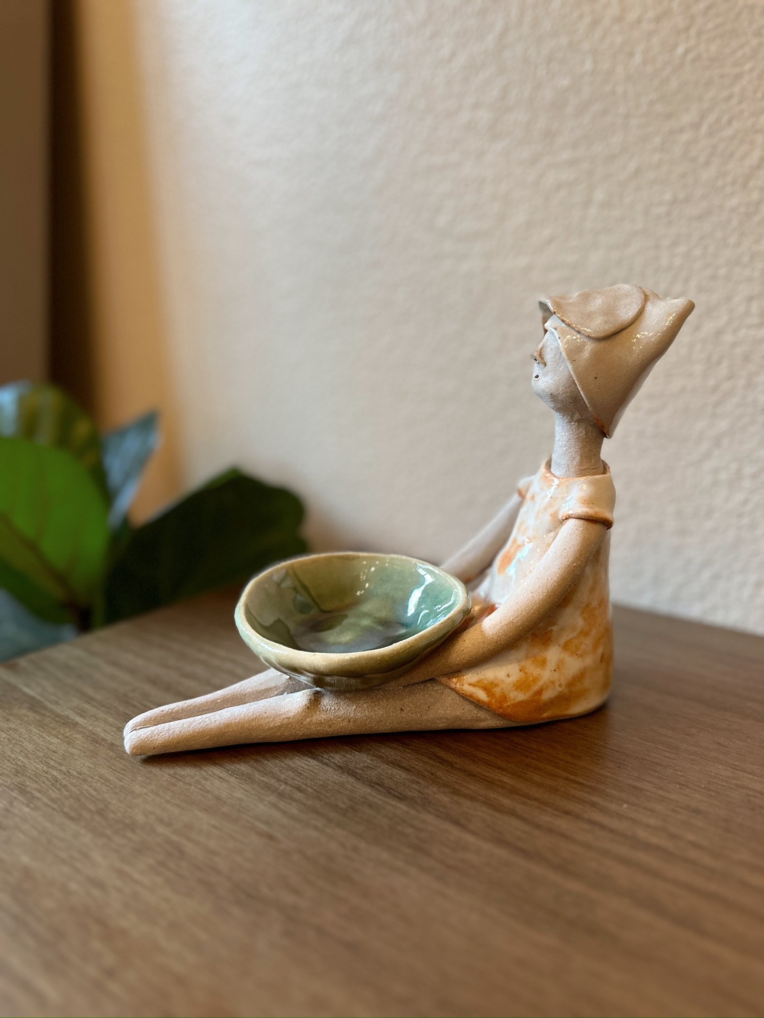 Glazed and Fired ceramic handmade sculpture of a lady holding a sage green bowl and sitting on a table. Image from the left.