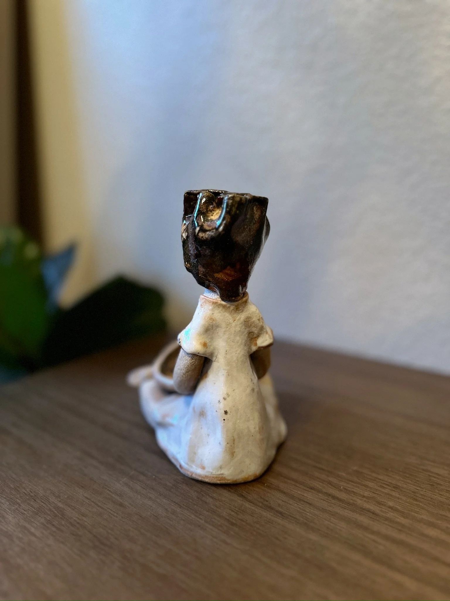 Glazed and Fired ceramic handmade sculpture of a lady with her hands in a prayer position, holding a small blue bowl with a bird on the edge, and sitting on a table. Image from the back.
