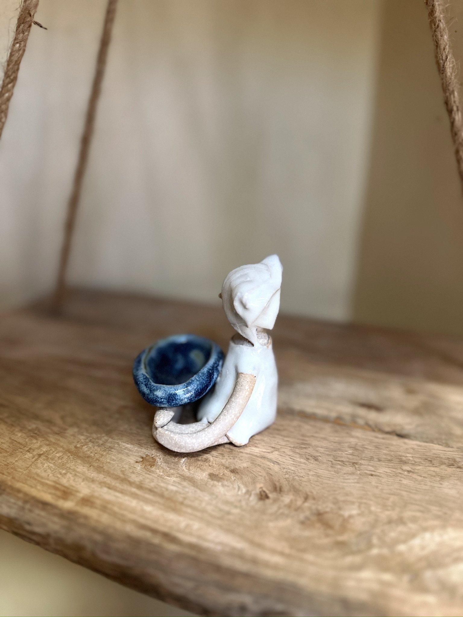 Glazed and Fired ceramic handmade sculpture of a lady with a small blue bowl and sitting on a swing. Image from the left.