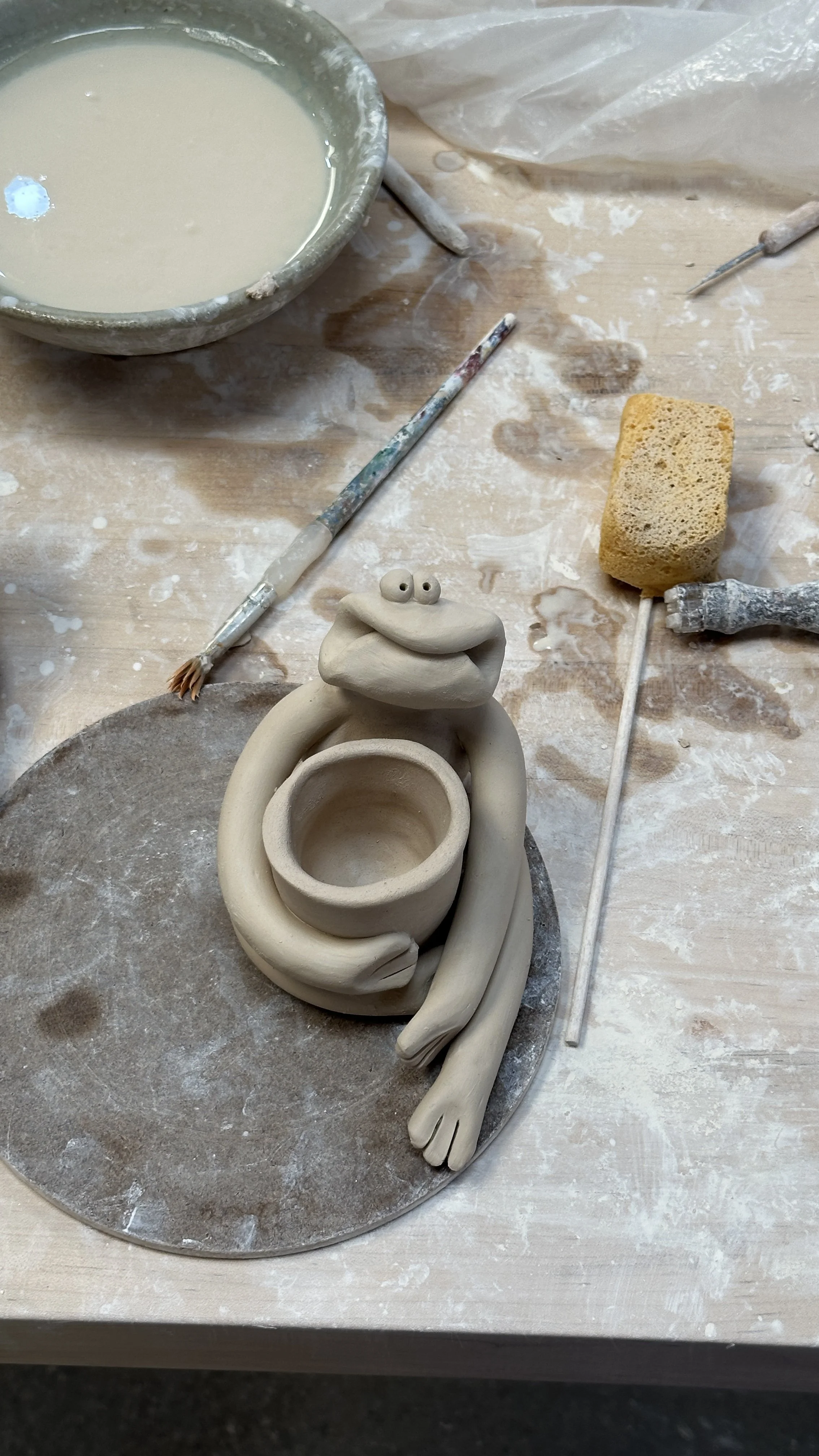 Making a sculpture of a frog holding a small vase. There are pottery tools around it showing the sculpture being in progress.