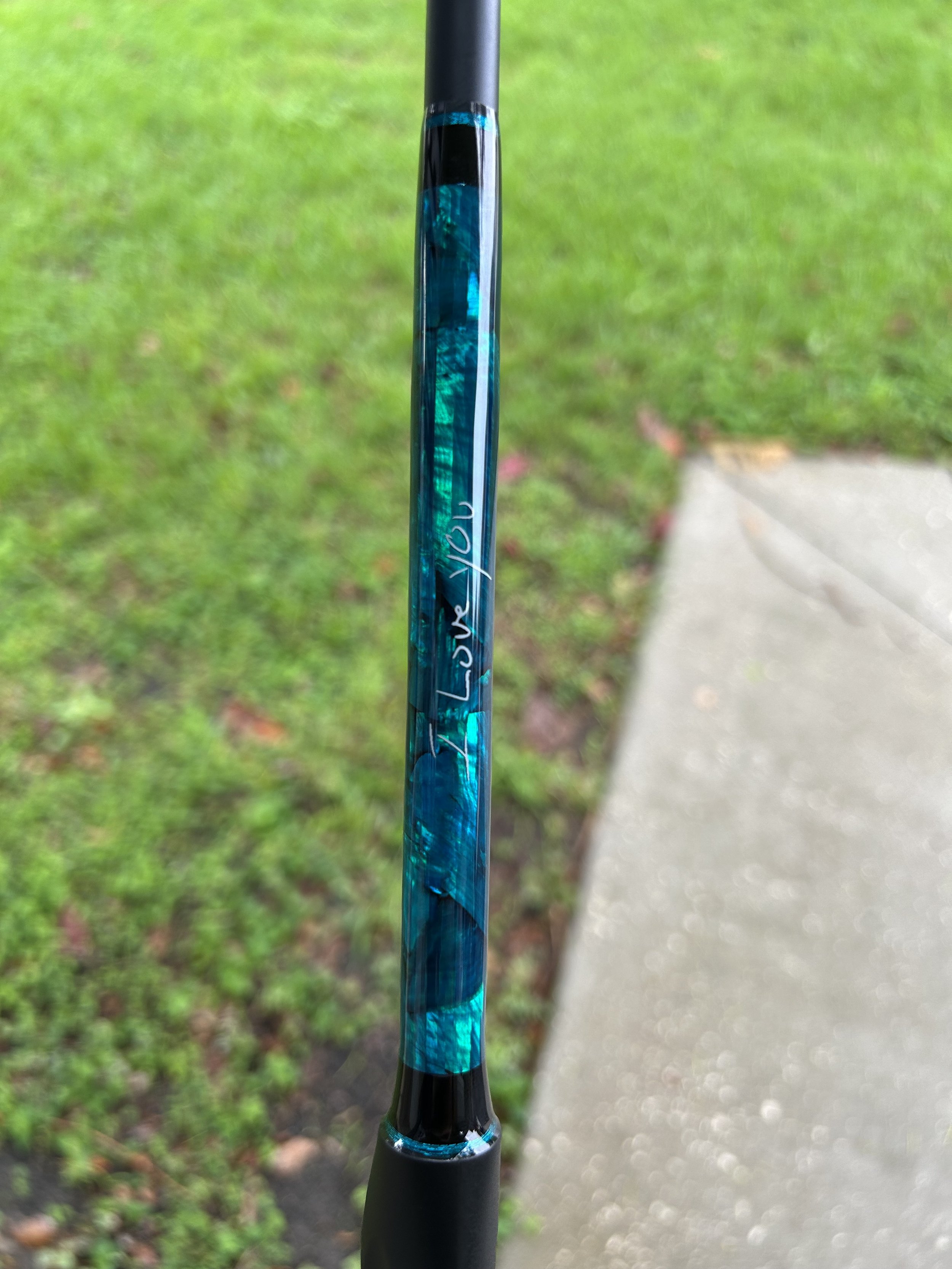 Close-up of a fishing rod with the word "Love" written on it, held outdoors on a rainy day with green grass and a concrete sidewalk in the background.
