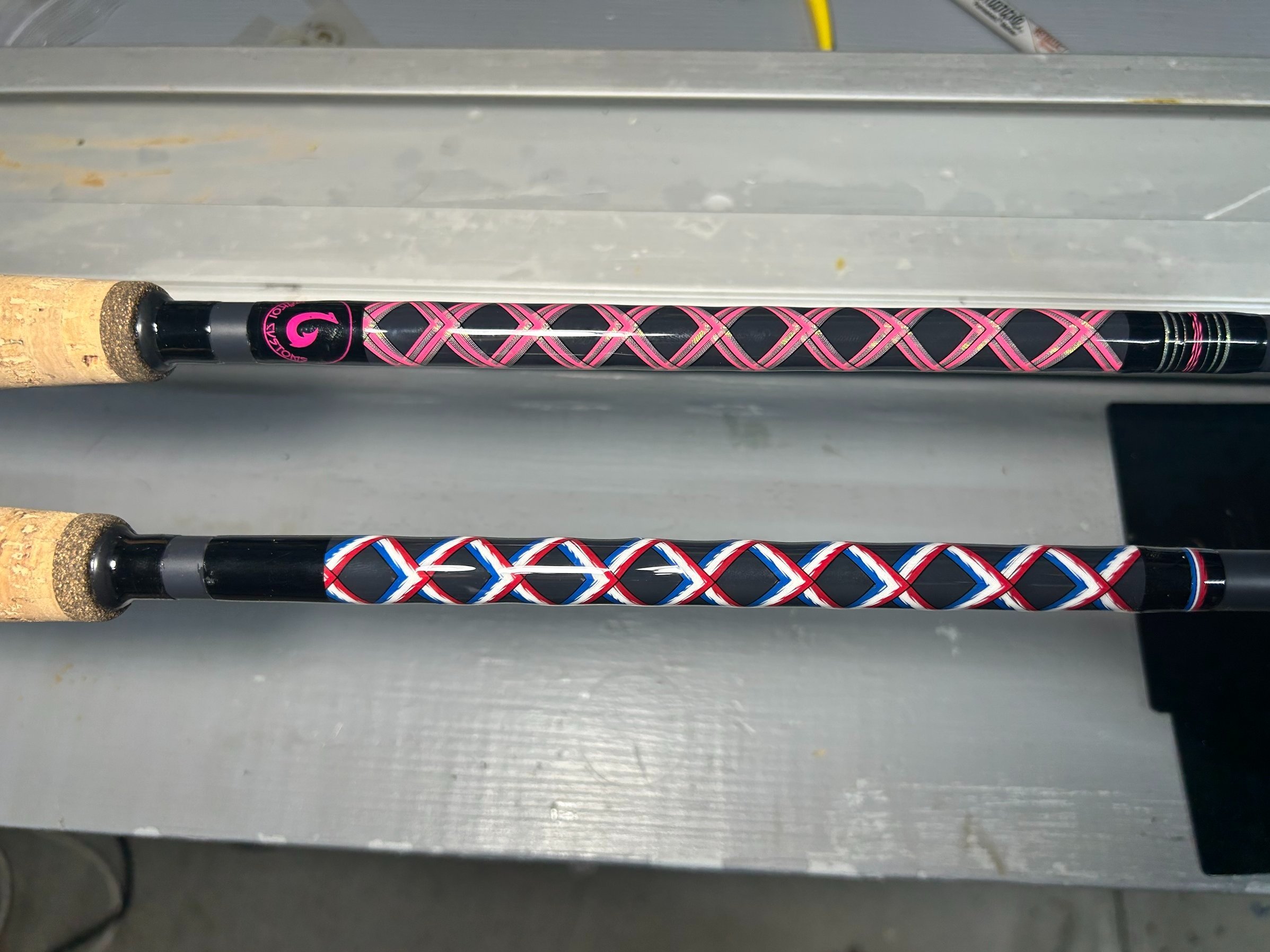 Two custom fishing rods with intricate wrapping designs, one in pink and black, the other in red, white, and blue, featuring cork handles.