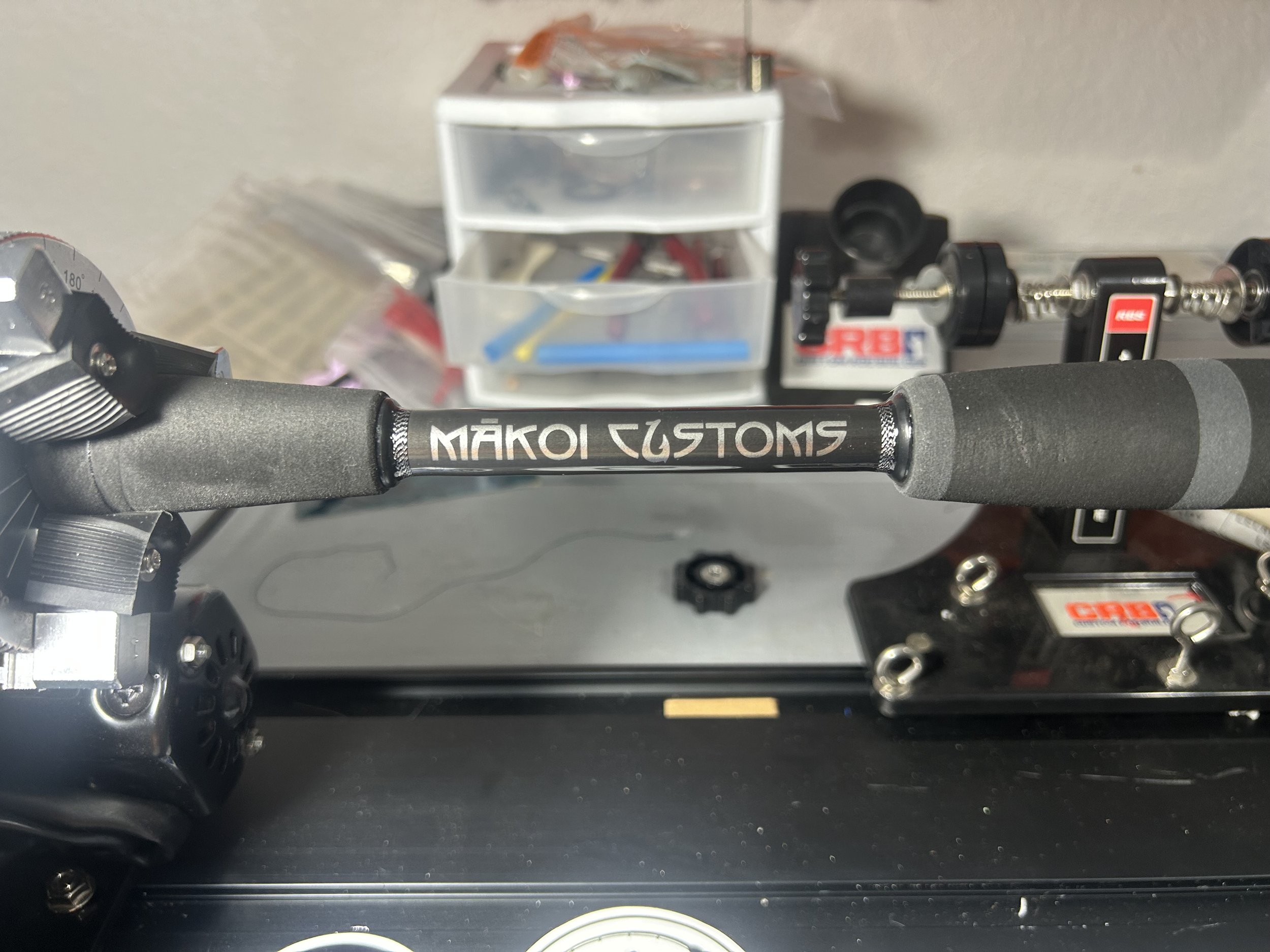 A Makoi Customs fishing rod mounted on a reel with a workbench in the background, containing a small plastic drawer, tools, and parts.