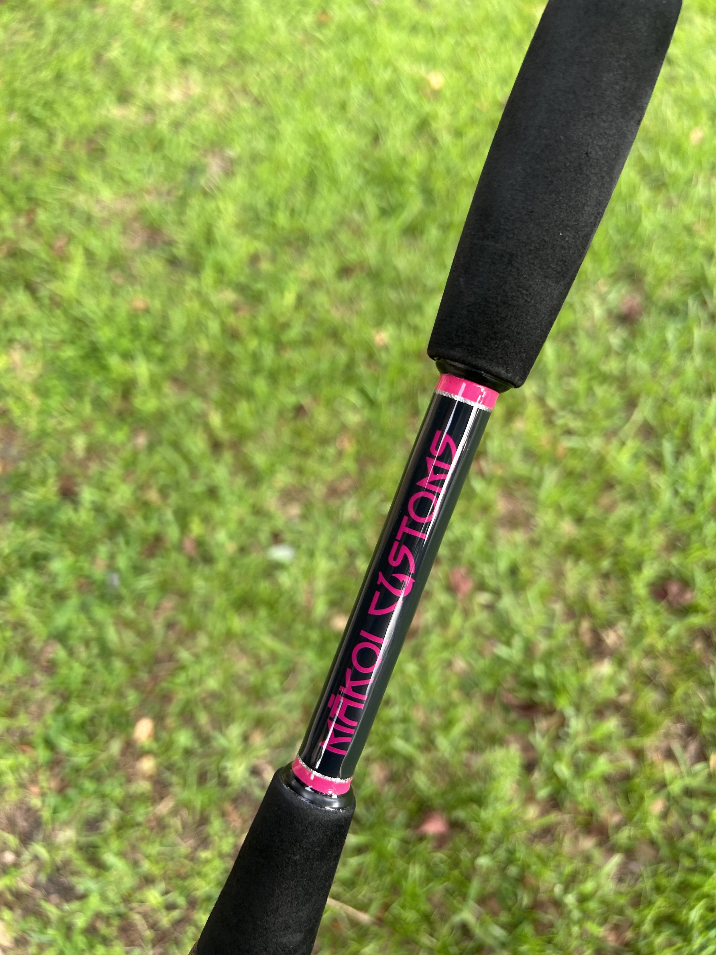 Close-up of a fishing rod with pink lettering and black foam grips, held above green grass.