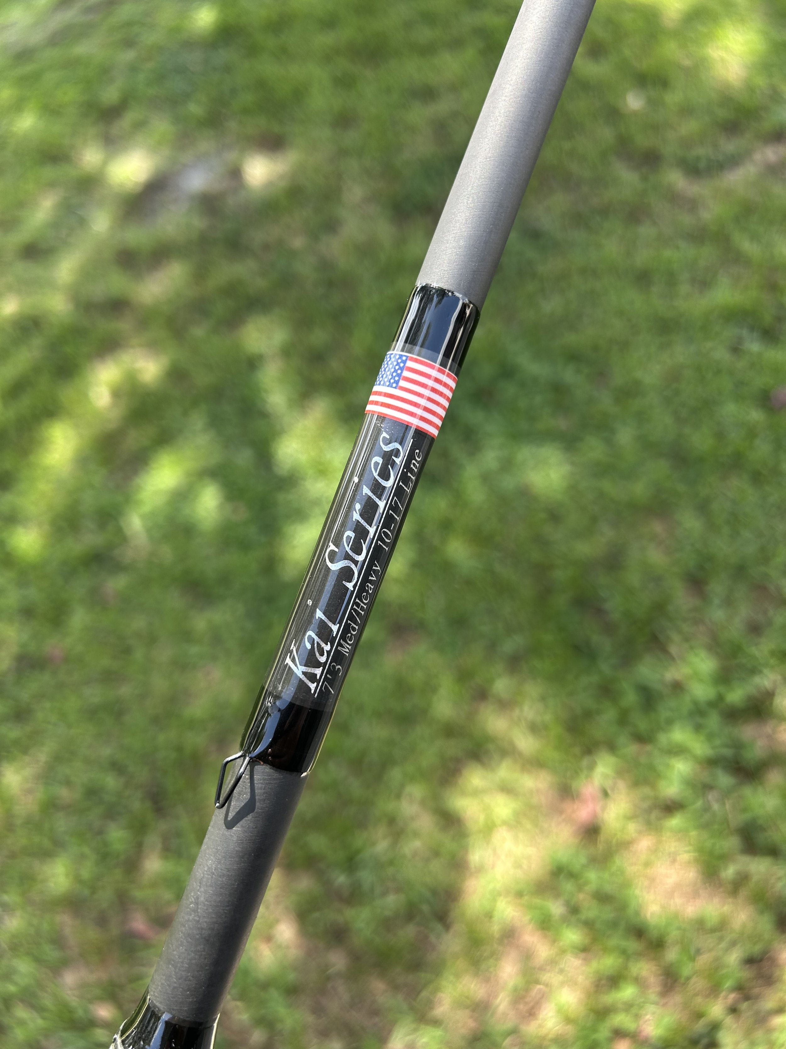 Close-up of a fishing rod labeled 'Kali Series 13', with an American flag sticker, held outdoors on grass.