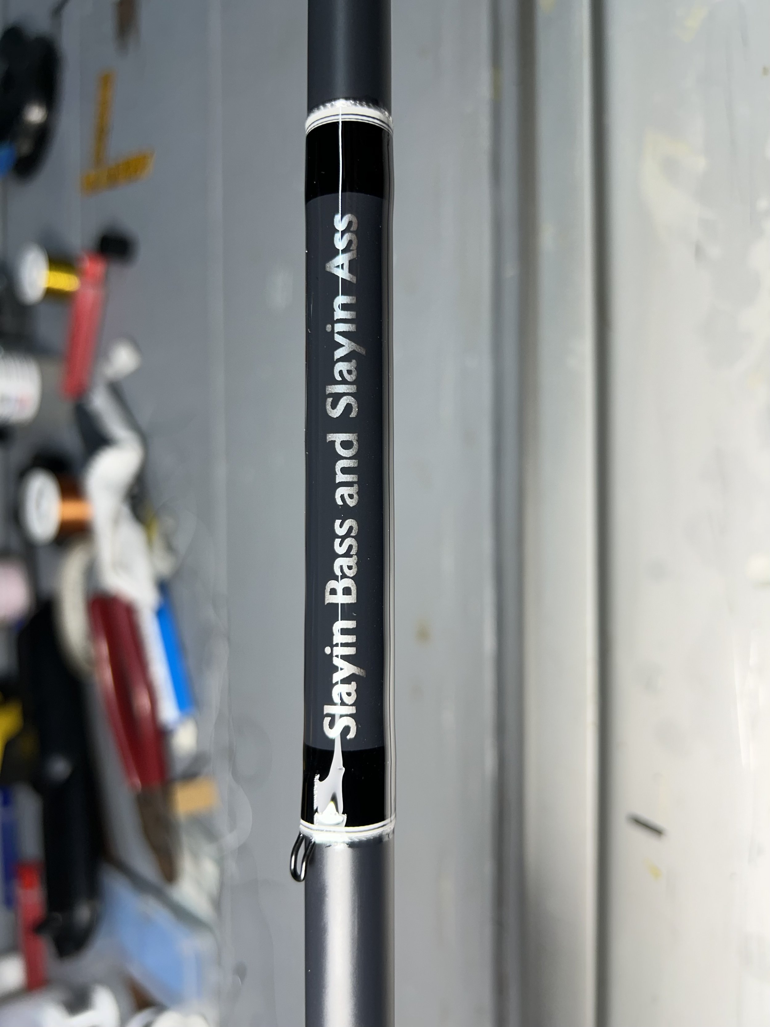 Fishing rod with text "Slayin Bass and Slayin Ass" printed on it, standing vertically against a blurred background in a storage area.