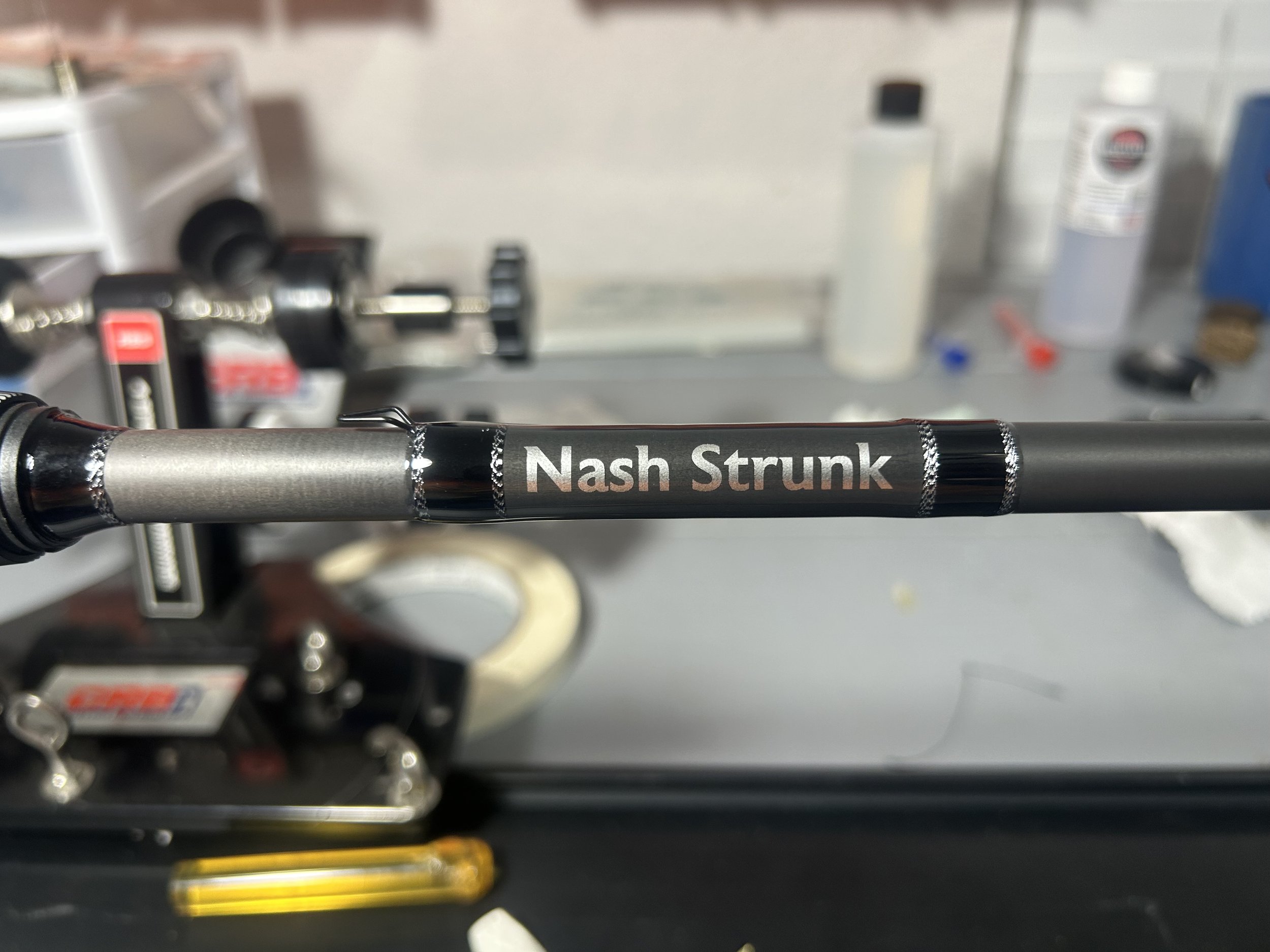 Close-up of a bicycle handlebar with a black wrap that reads 'Nash Strunk' in white letters. The background shows a workbench with various tools and bottles, slightly out of focus.