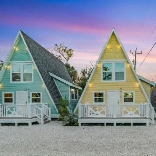 Two colorful A-frame houses with white railings and string lights, set against a sunset sky.