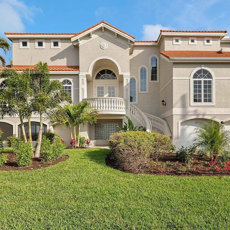 Front view of a large, elegant beige house with a red tile roof, arched windows, and a balcony with white balustrades, surrounded by lush green grass and tropical plants.