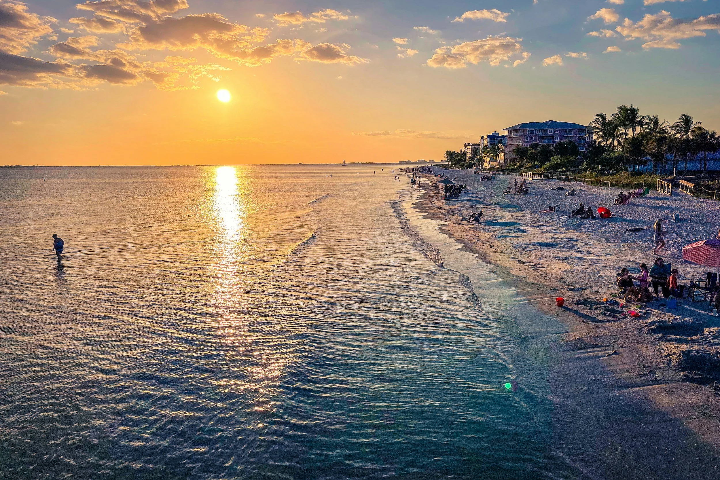 A sunset over a beach with people relaxing and swimming in the water, and buildings and palm trees lining the shoreline.