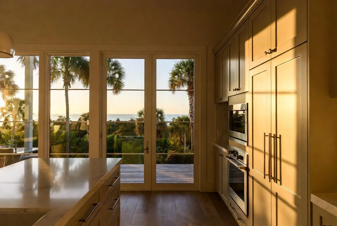 Kitchen with large glass doors opening to an outdoor deck, overlooking palm trees and the ocean during sunset.