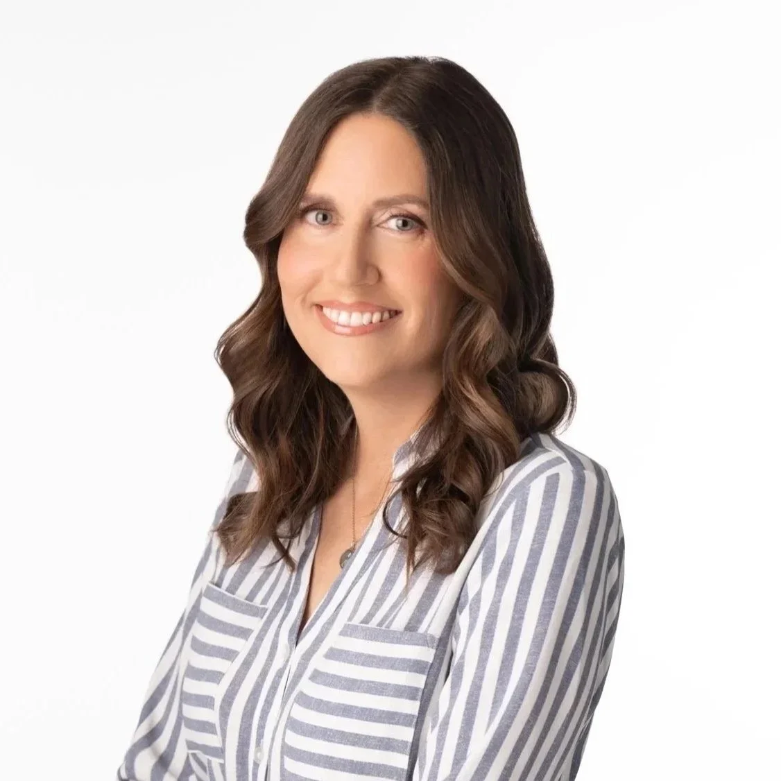 A woman with long brown hair styled in loose waves, wearing a white and gray striped shirt, smiling against a white background.