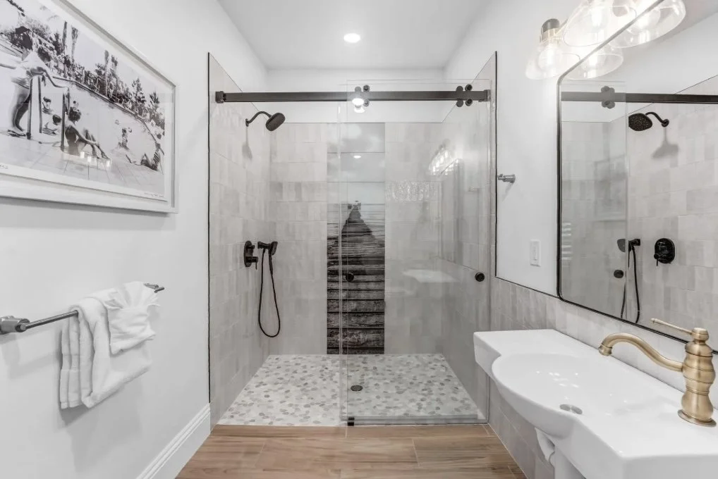 A modern bathroom with a walk-in glass shower, white walls, a large mirror, a white sink with gold fixtures, and a towel rack with towels.