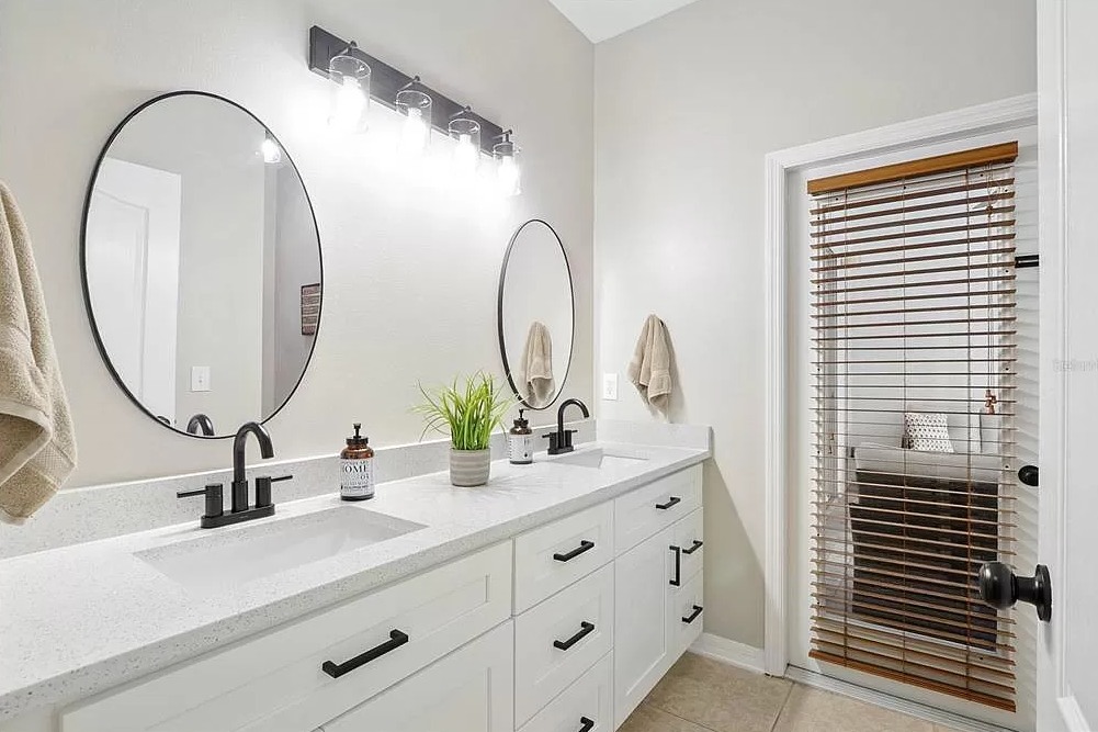 A modern bathroom with a large white double vanity, black hardware, two oval mirrors, and a window with wooden blinds. There are two beige towels hanging on the wall, and a potted green plant on the countertop. Overhead lighting features four exposed