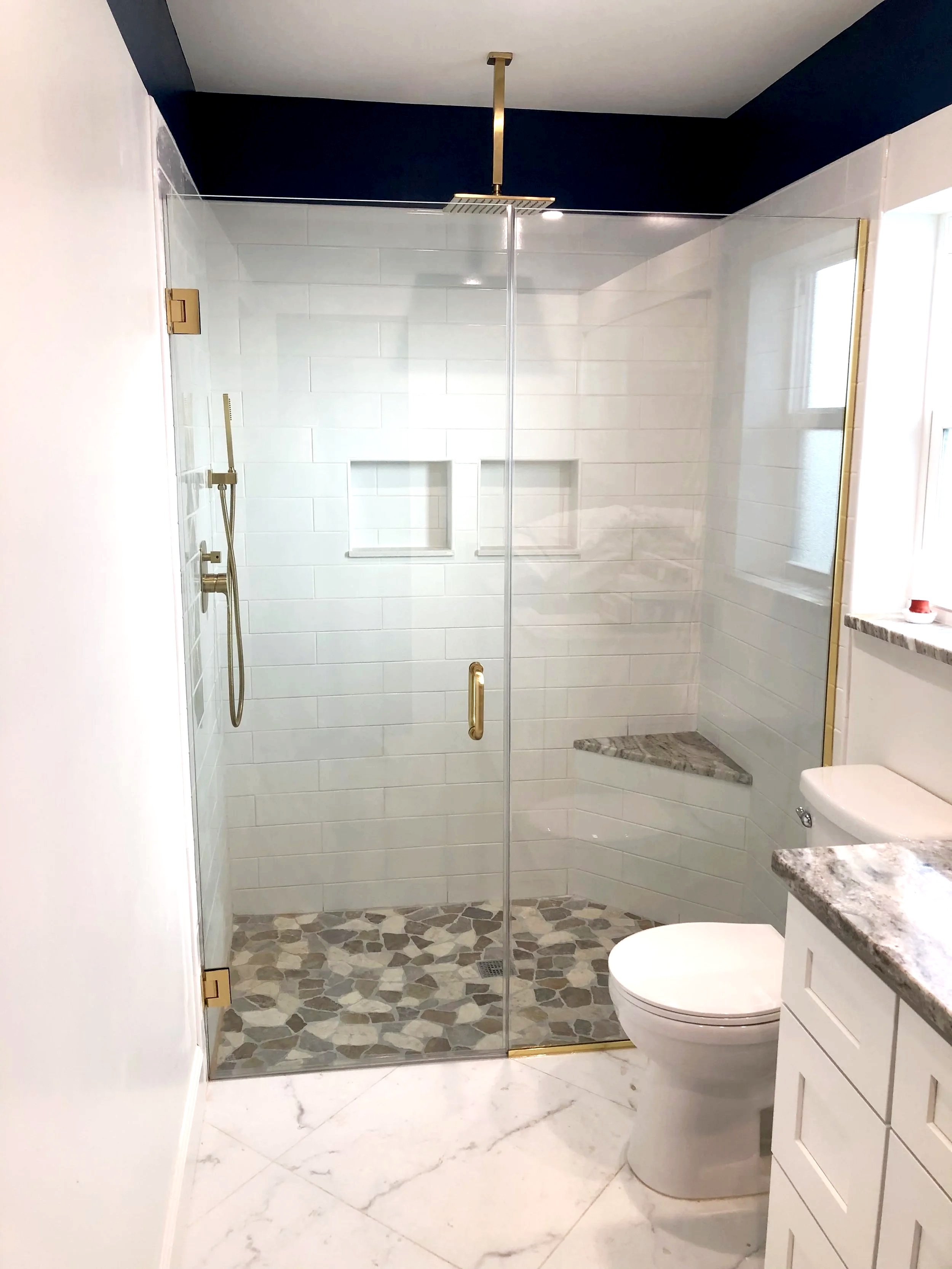 Modern bathroom with walk-in shower featuring glass walls, gold fixtures, and marble floor tiles. The bathroom includes a toilet and a white vanity with a granite countertop.