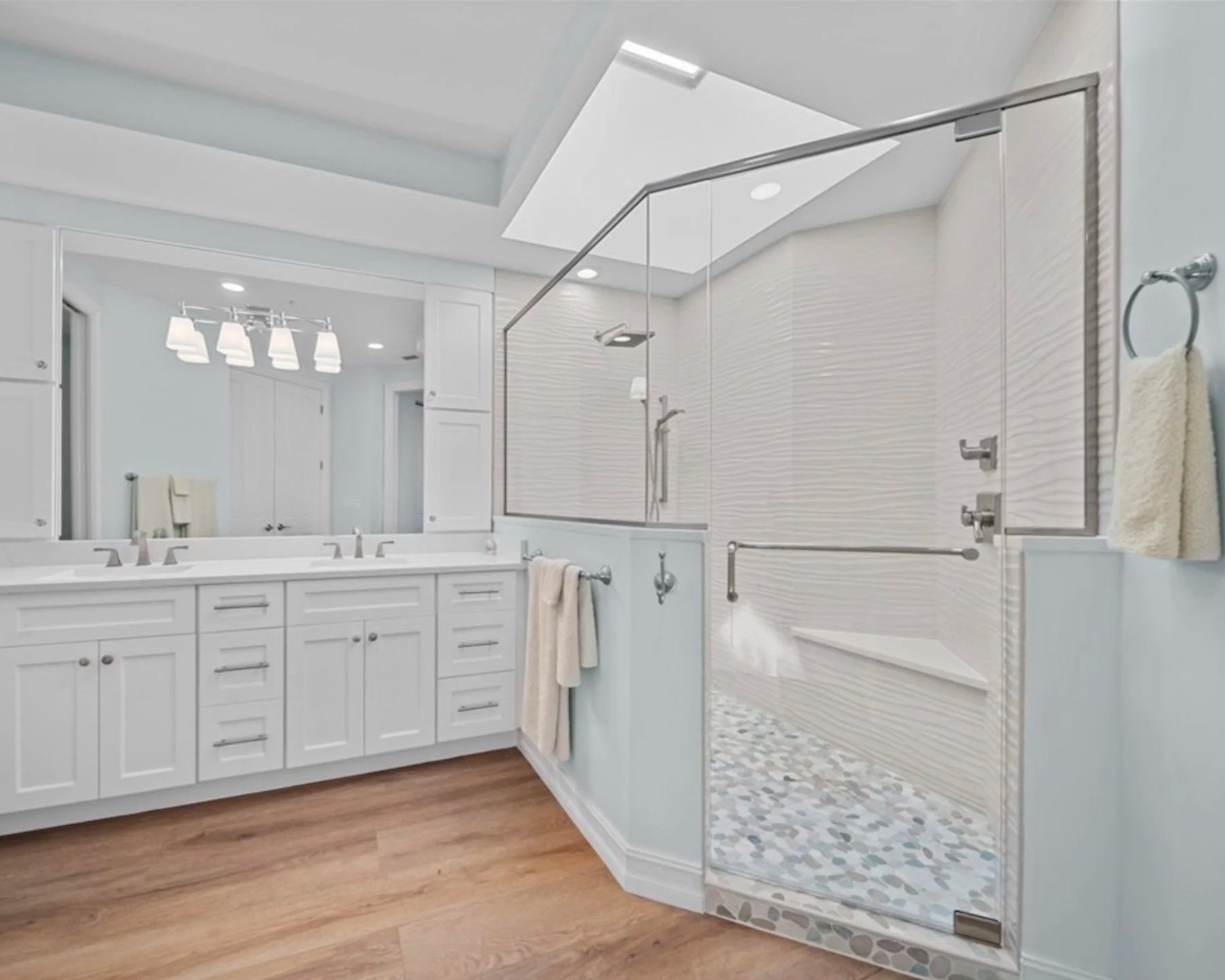 Modern white bathroom with double vanity, large mirror, and walk-in shower with glass door and pebble floor.