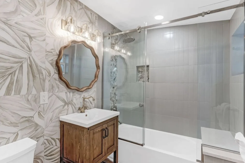 Modern bathroom with a leaf-patterned wallpaper, a wooden vanity with a white sink, and a glass-enclosed shower with gray tiles.