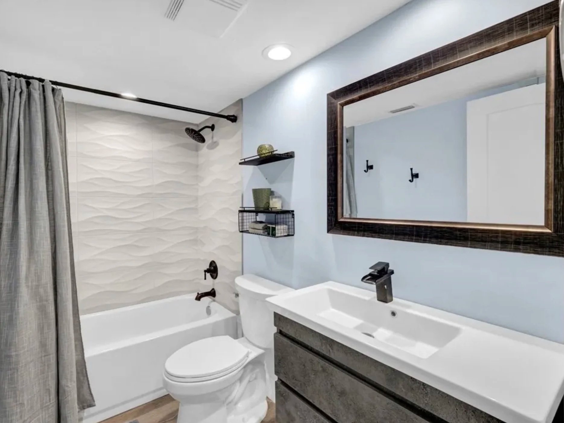 Modern bathroom with a bathtub and shower, gray textured wall, white and black fixtures, a large mirror with a dark wood frame, a white sink with a black faucet, and gray and black storage cabinet.