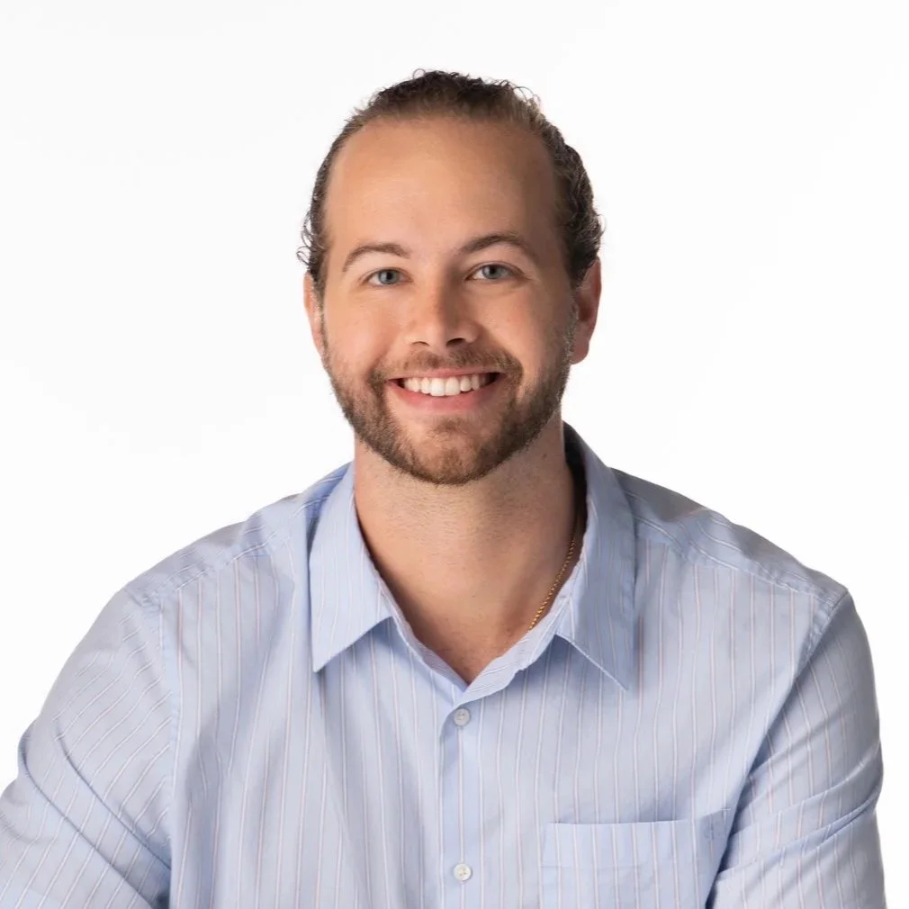 A smiling man with short, light brown hair and a beard, wearing a light blue, striped dress shirt, against a white background.