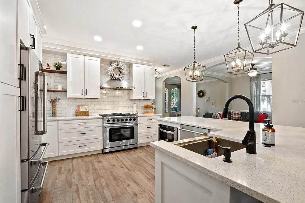 Modern kitchen with white cabinets, stainless steel appliances, a large island with a sink, and pendant lighting fixtures. The background features a living room with a sofa and decorative wreaths.