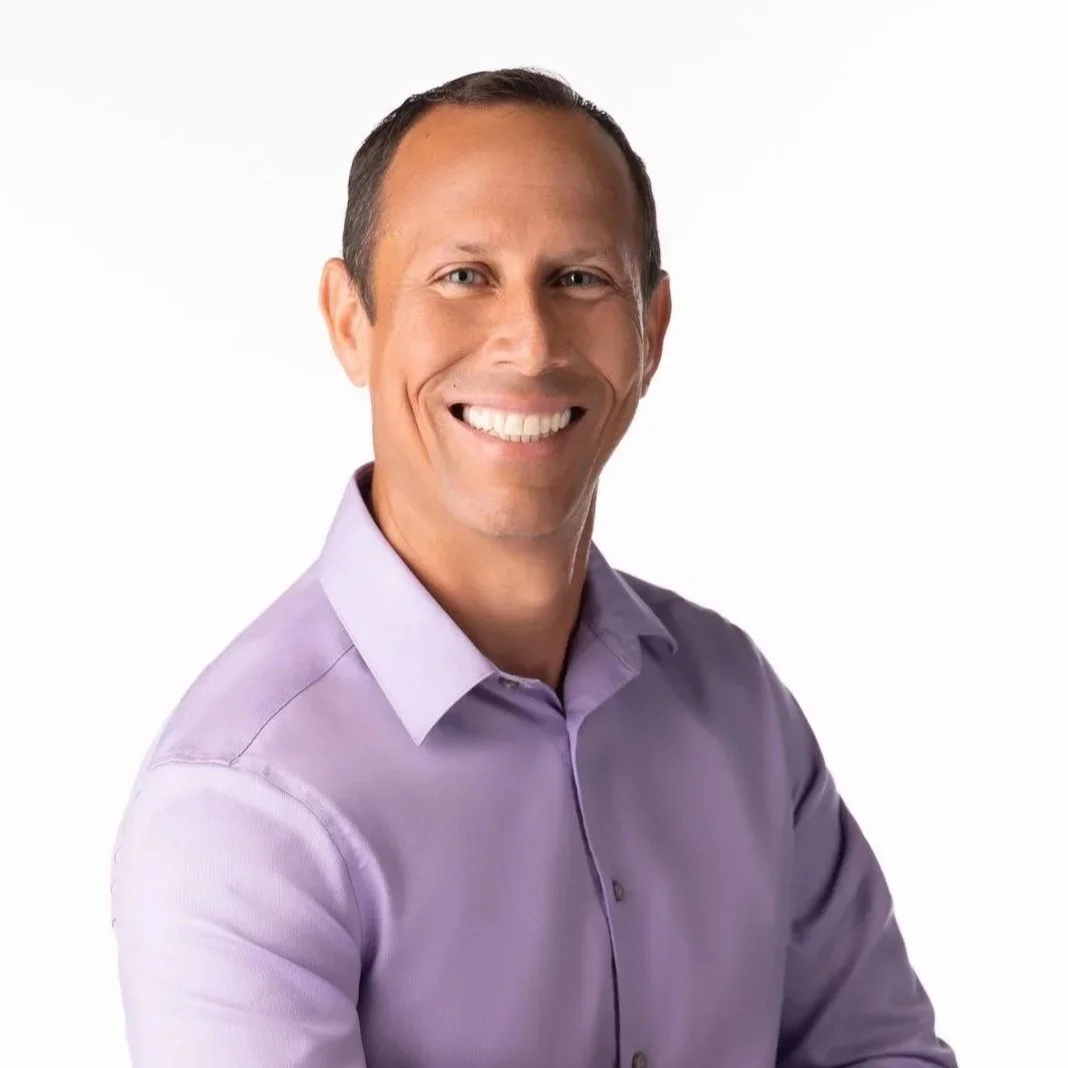 A smiling man wearing a light purple dress shirt against a white background.