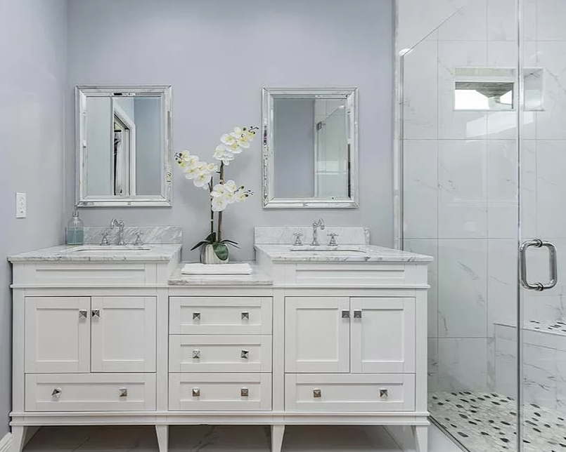 A white bathroom vanity with two sinks, two mirrors, and a decorative orchid in the middle, with a glass shower enclosure on the right.
