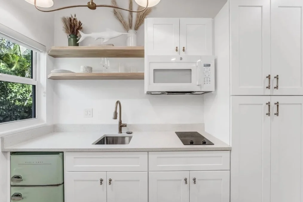 Modern small kitchen with white cabinets, a sink with a bronze faucet, a microwave oven above the stove, two floating wooden shelves with decorative items, a large window with greenery outside, and a light green mini refrigerator.