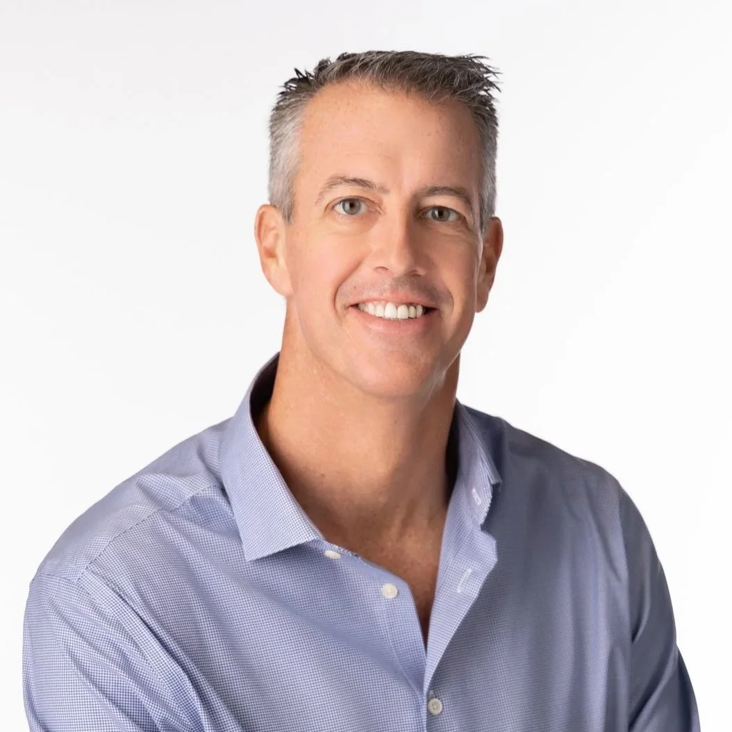 A smiling man with short, graying hair wearing a light purple dress shirt, against a plain white background.