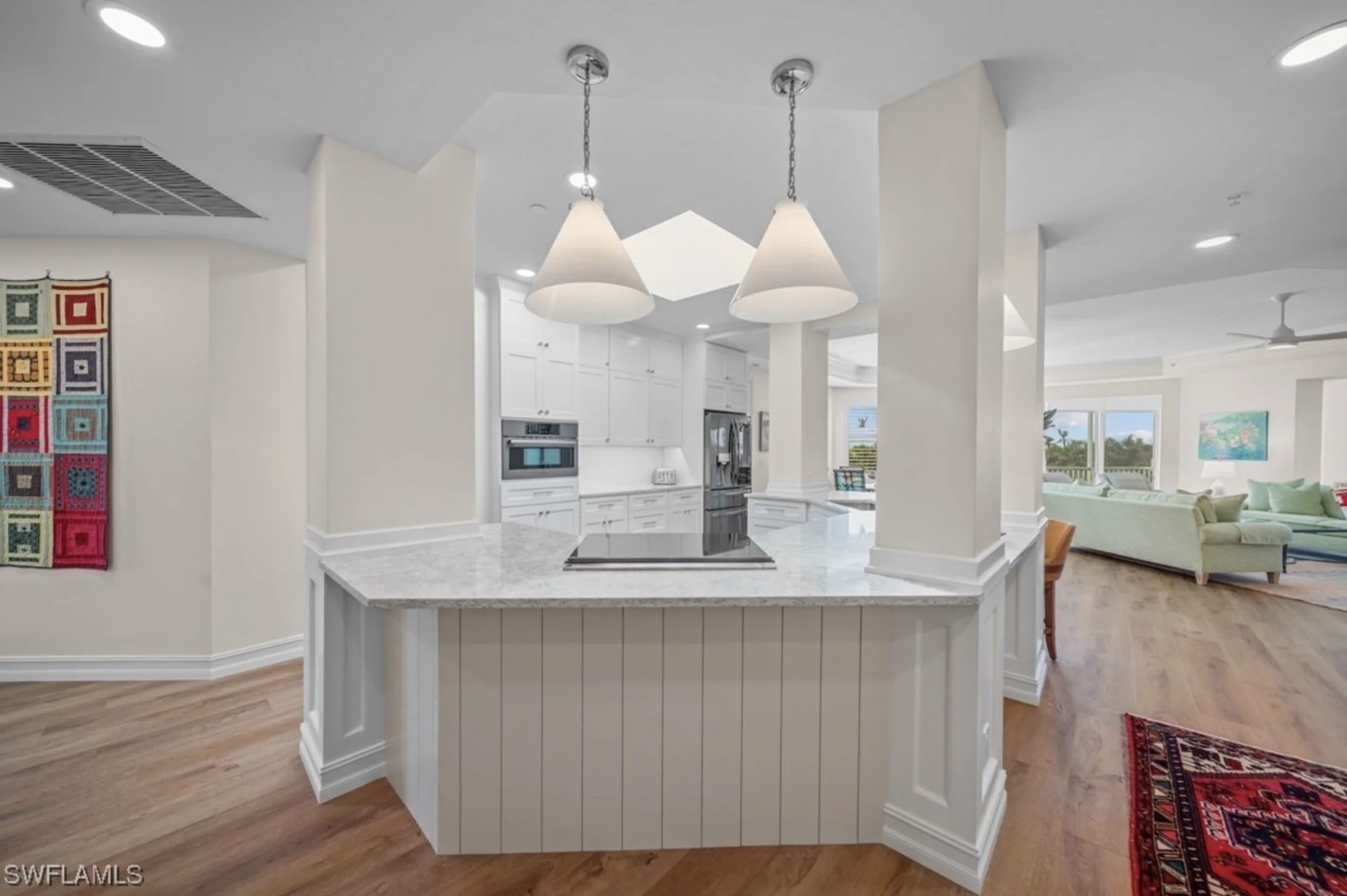 A modern kitchen with white cabinets, granite countertops, and a kitchen island. Two cone pendant lights hang above the island. In the background, part of a living room with a green sofa and windows is visible.