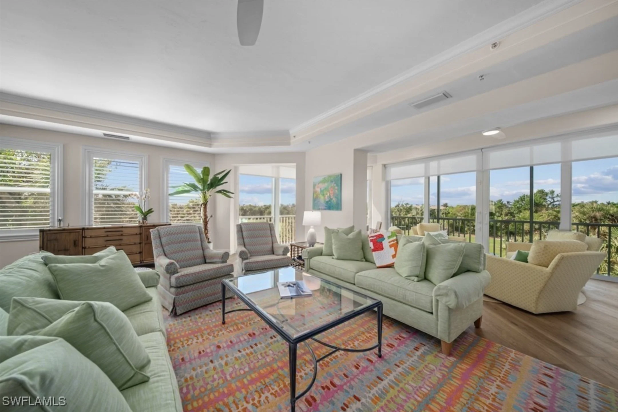 Living room with light green couches, patterned armchairs, a glass coffee table, colorful area rug, potted plant, large windows with blinds, and a view of greenery outside.