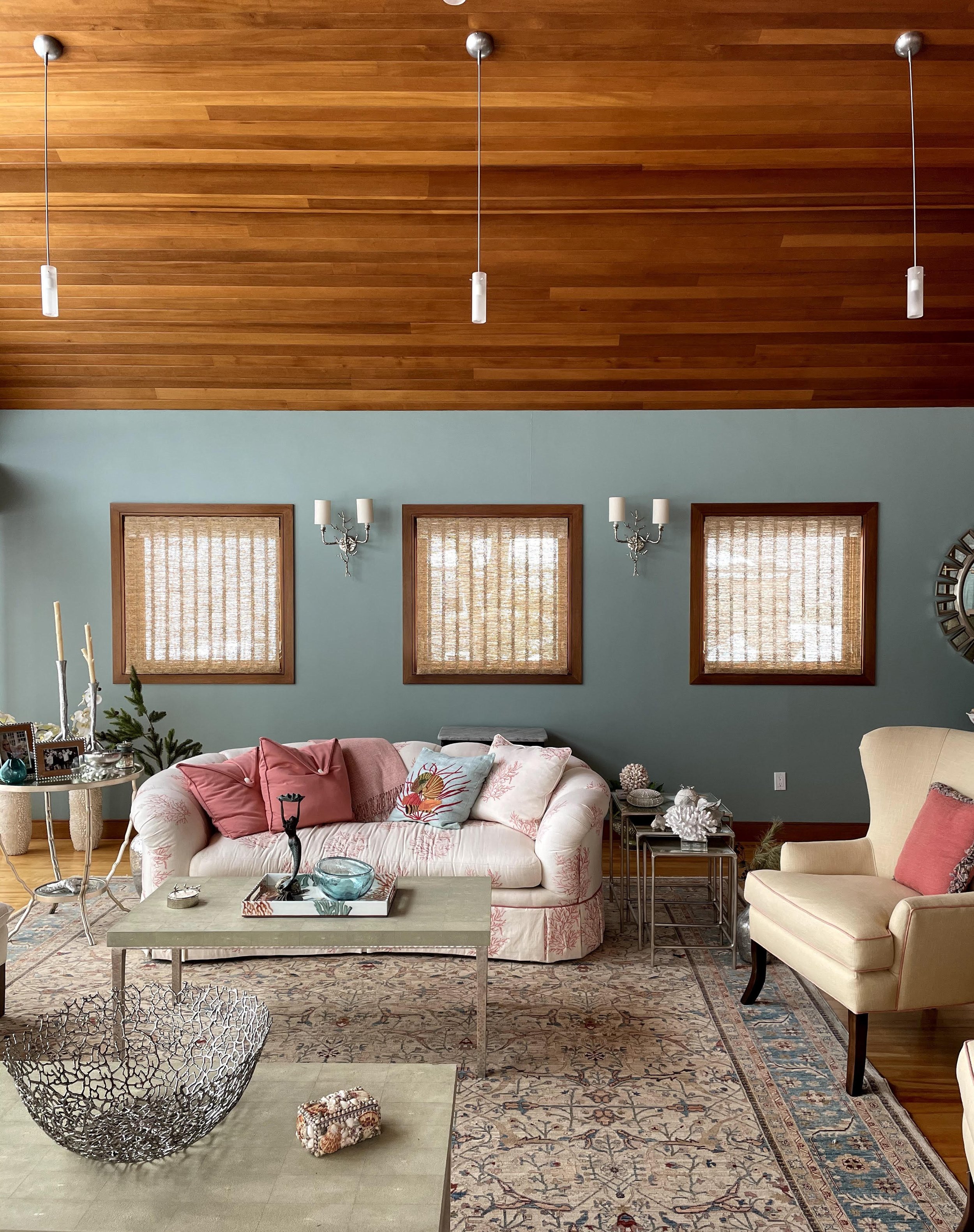 Cozy living room with a white sofa with pink cushions, patterned armchair, wooden ceiling, light blue walls, and decorative wall sconces. A patterned rug covers the floor, and a metal decorative bowl is on the coffee table.