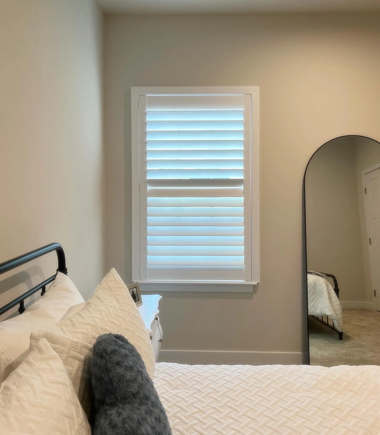 Minimalist bedroom with white bedding, a metal headboard, a tall mirror, and a window with white blinds.