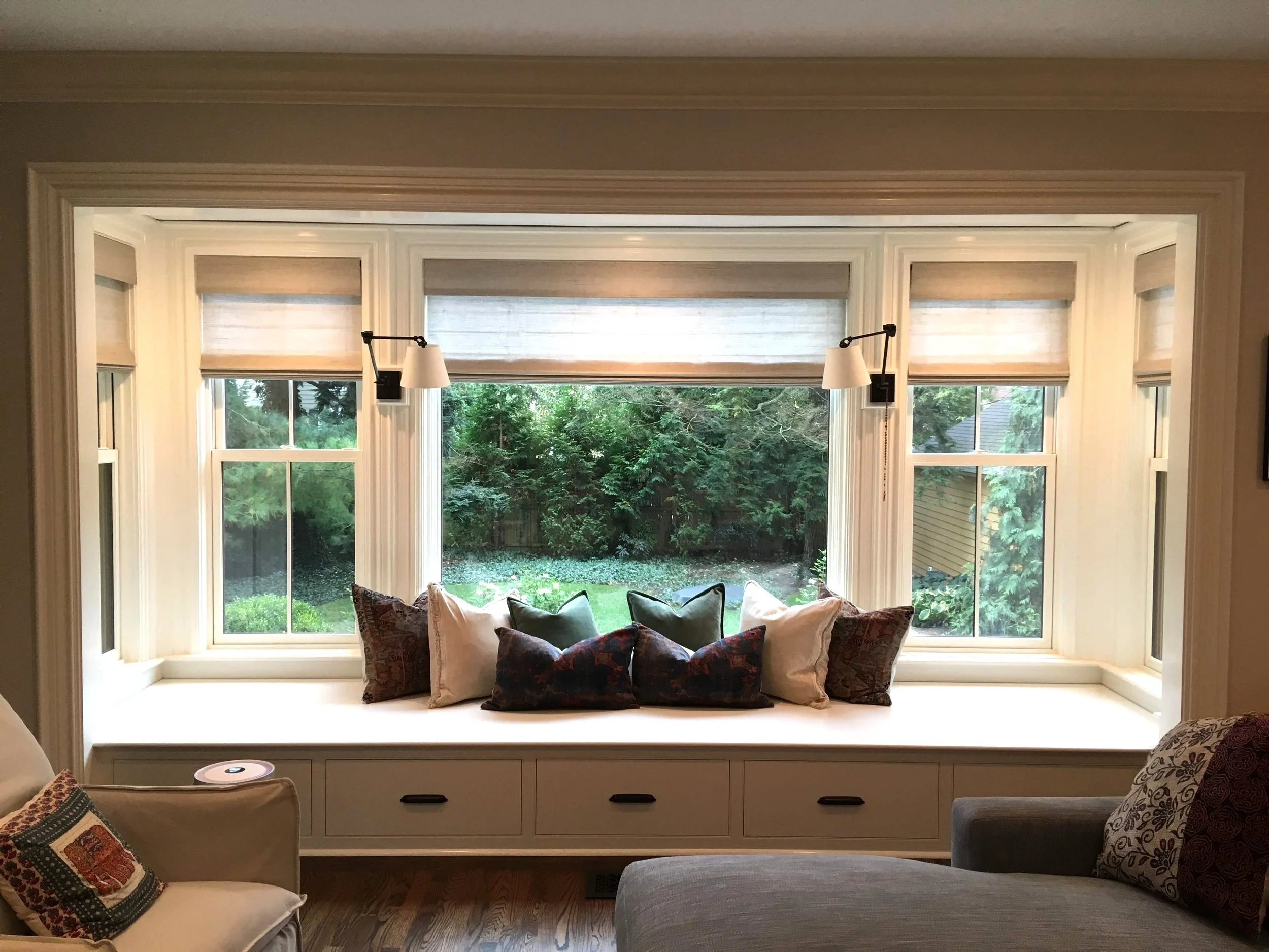 Cozy window seat with cushions, surrounded by large windows offering a view of a lush garden, with a nearby armchair and wood flooring.