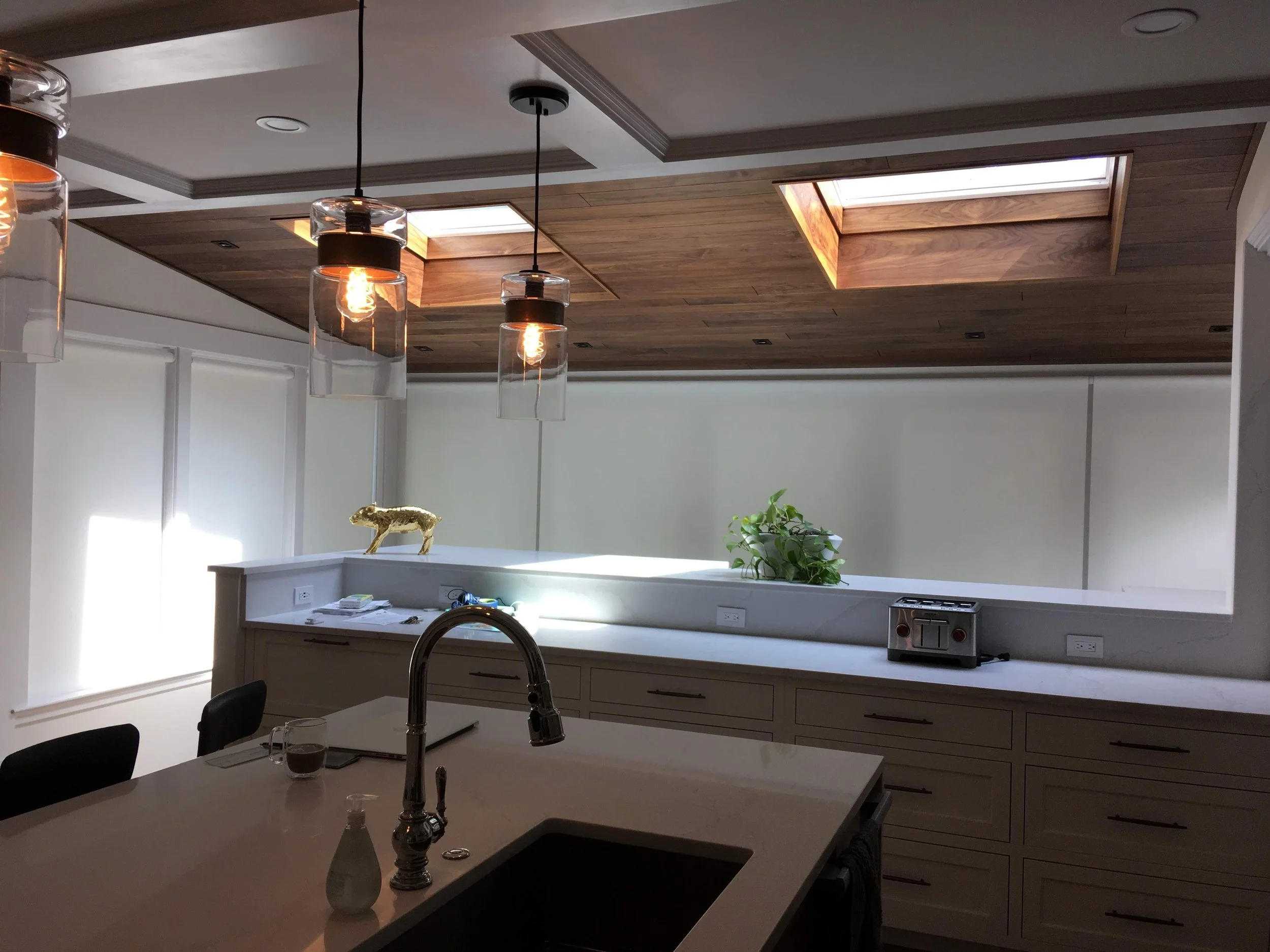 Modern kitchen with pendant lights, skylights, quartz countertops, sink, faucet, toaster, decorative golden animal figurine, and potted plant.