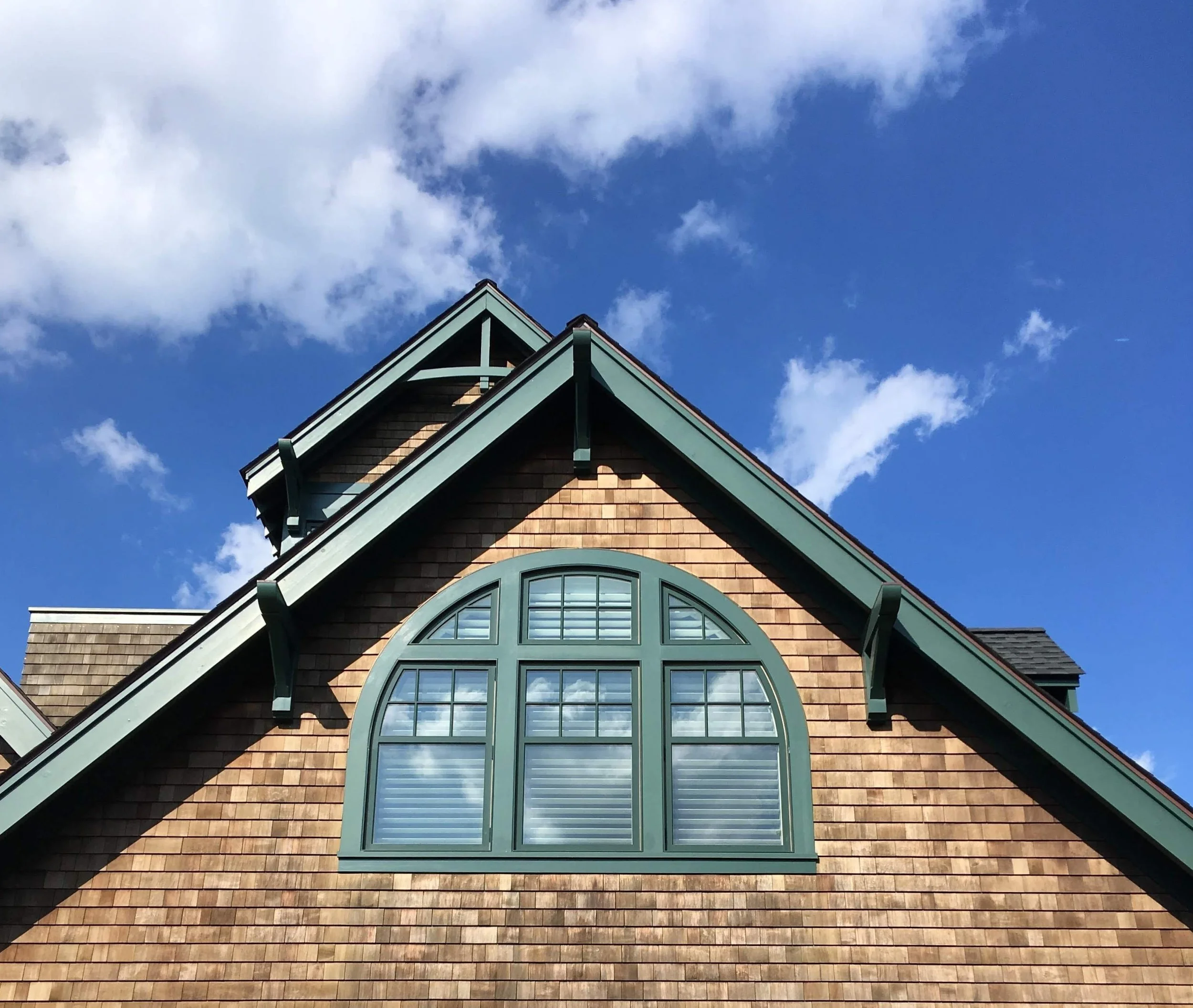 Gable roof with brown shingles and arched window against blue sky with clouds.