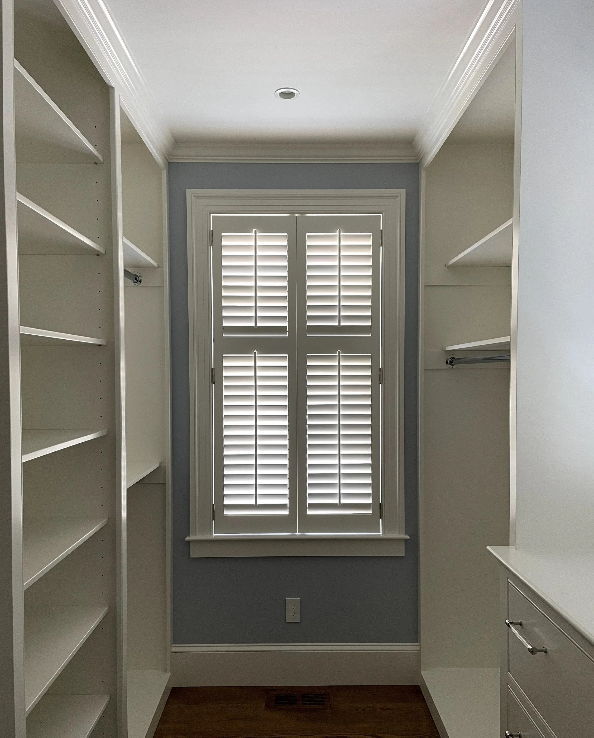Walk-in closet with white shelves, hanging rods, and shutters on window.