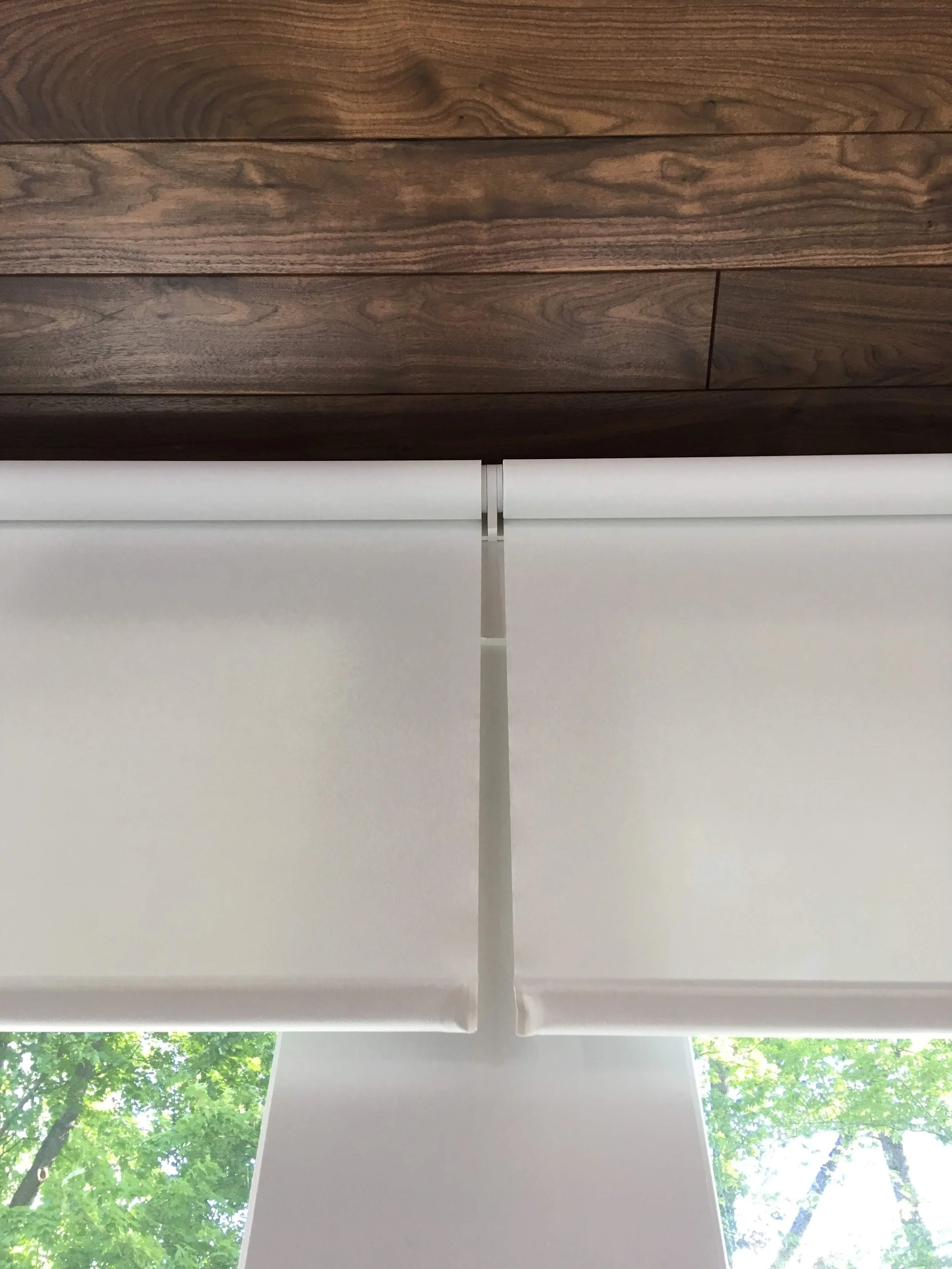 White roller blinds partially lowered in front of a window with a view of green trees outside, beneath a wooden ceiling.