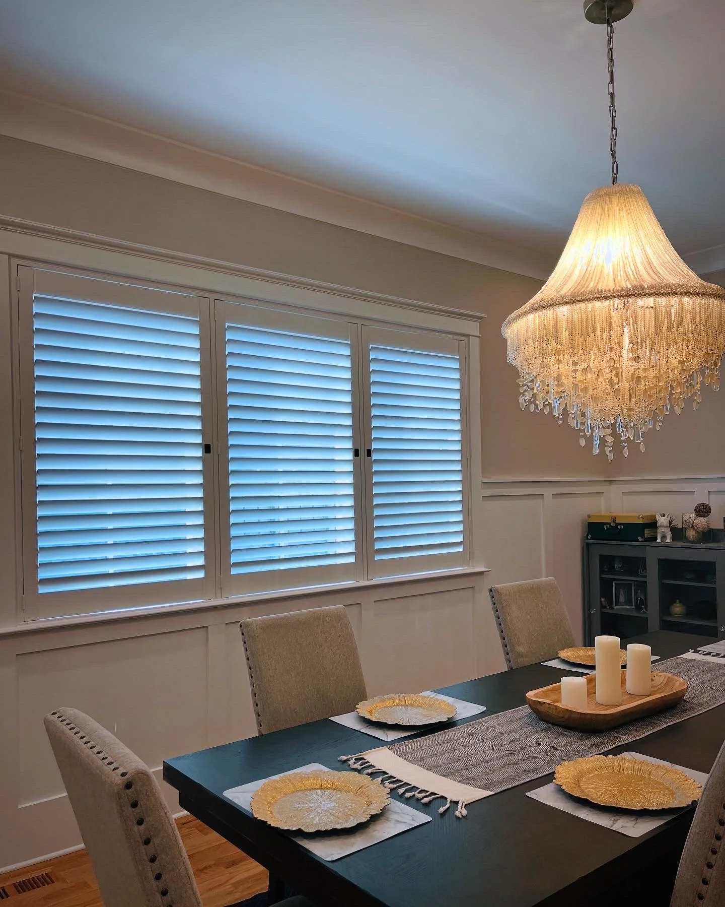 Elegant dining room with a dark wood table set for four, featuring textured gold plates and a gray runner. A wooden tray with candles is in the center. The room is lit by a large, ornate chandelier and has windows with white shutters.