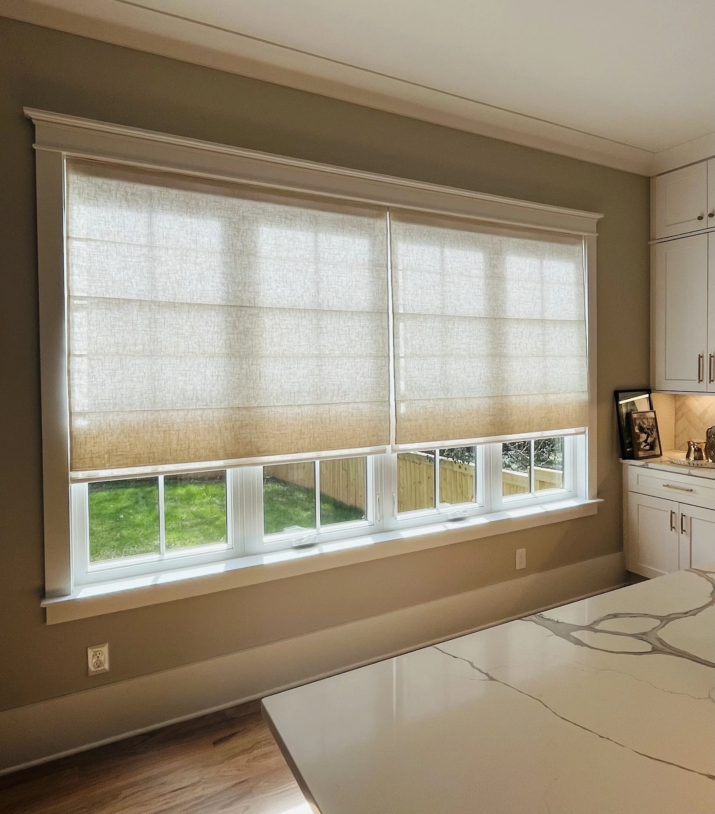 Kitchen interior with large window covered by beige Roman shades, a marble countertop, and light wood flooring.