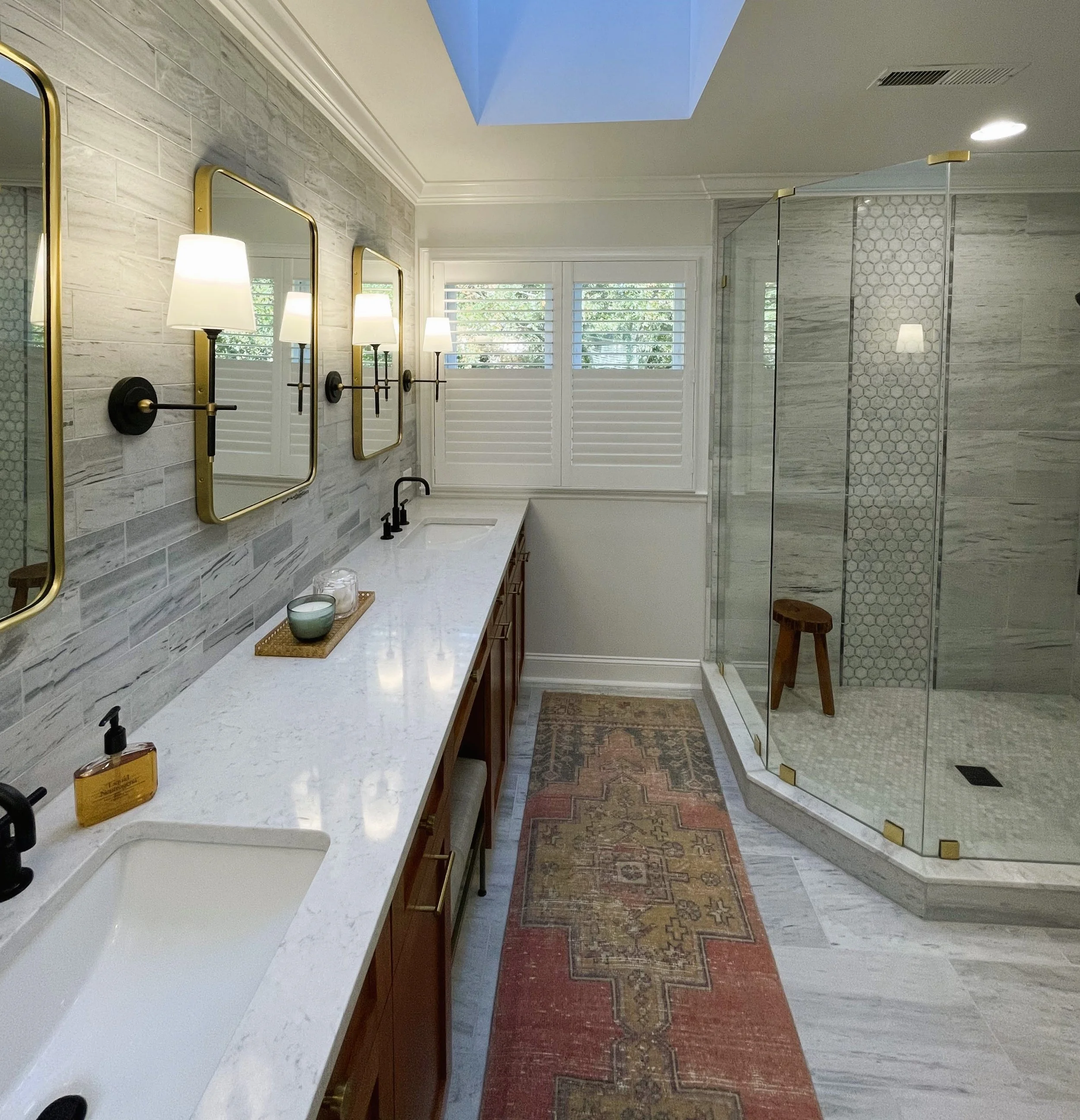 Elegant bathroom with a double sink vanity and marble countertop, featuring gold-framed mirrors and wall sconces. A colorful runner rug is on the floor next to a large glass shower with hexagonal tile design. A skylight and window provide natural lig