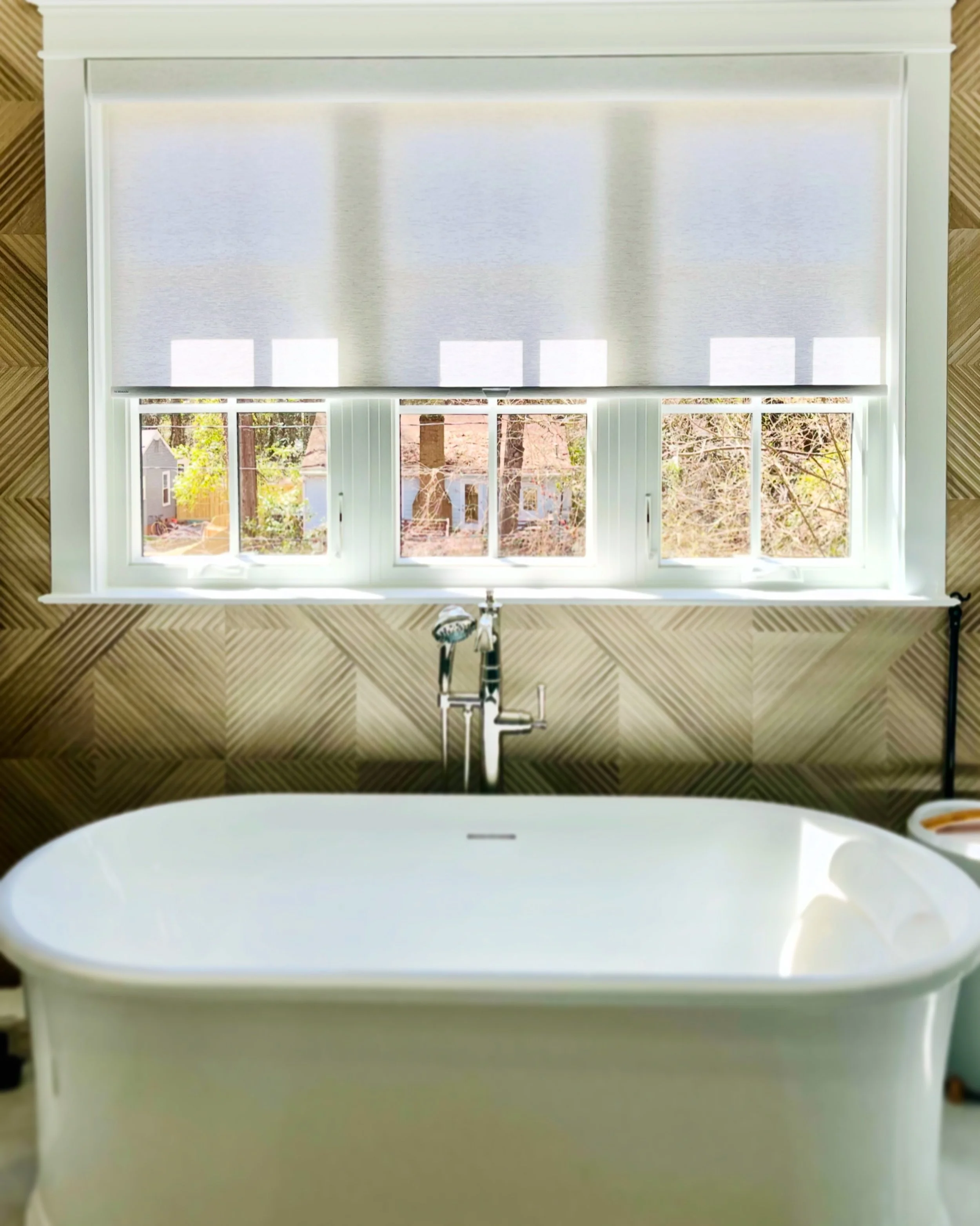 White freestanding bathtub with a modern faucet in front of a large window with partially drawn blinds, featuring a geometric textured wall background.