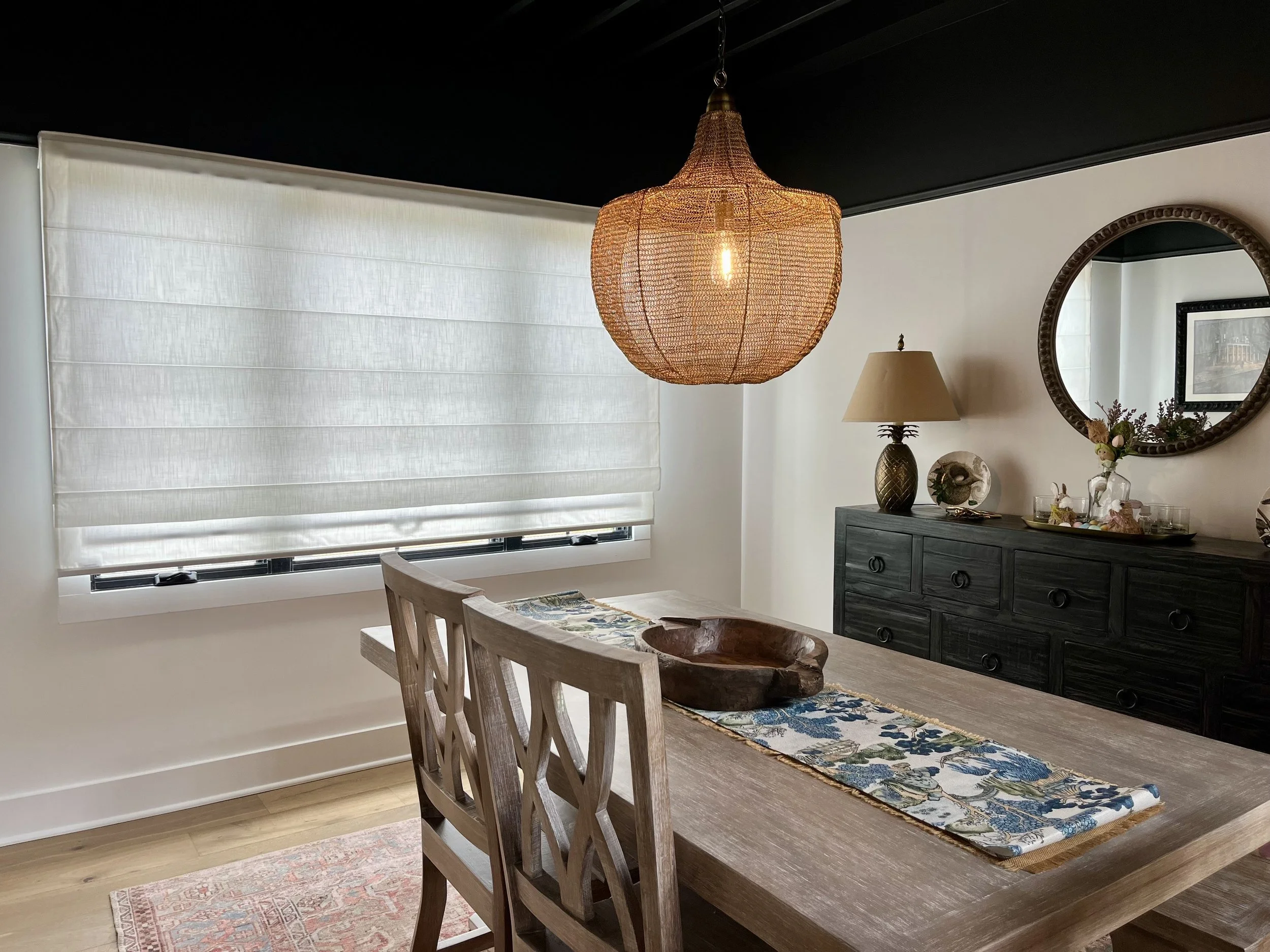 Dining room with large woven pendant light, wooden table with floral runner, and black dresser with decorative items and mirror.