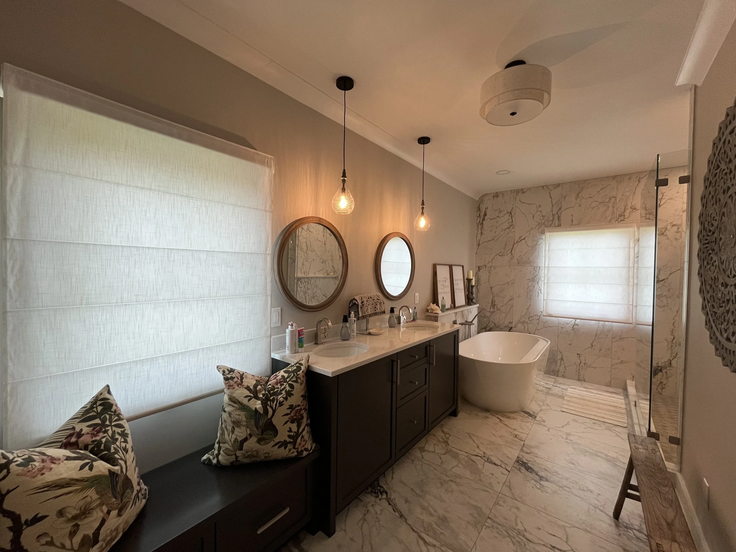 Luxurious bathroom with marble floors and walls, a freestanding white bathtub, double sinks with round mirrors, pendant lighting, a cushioned bench, and decorative elements.