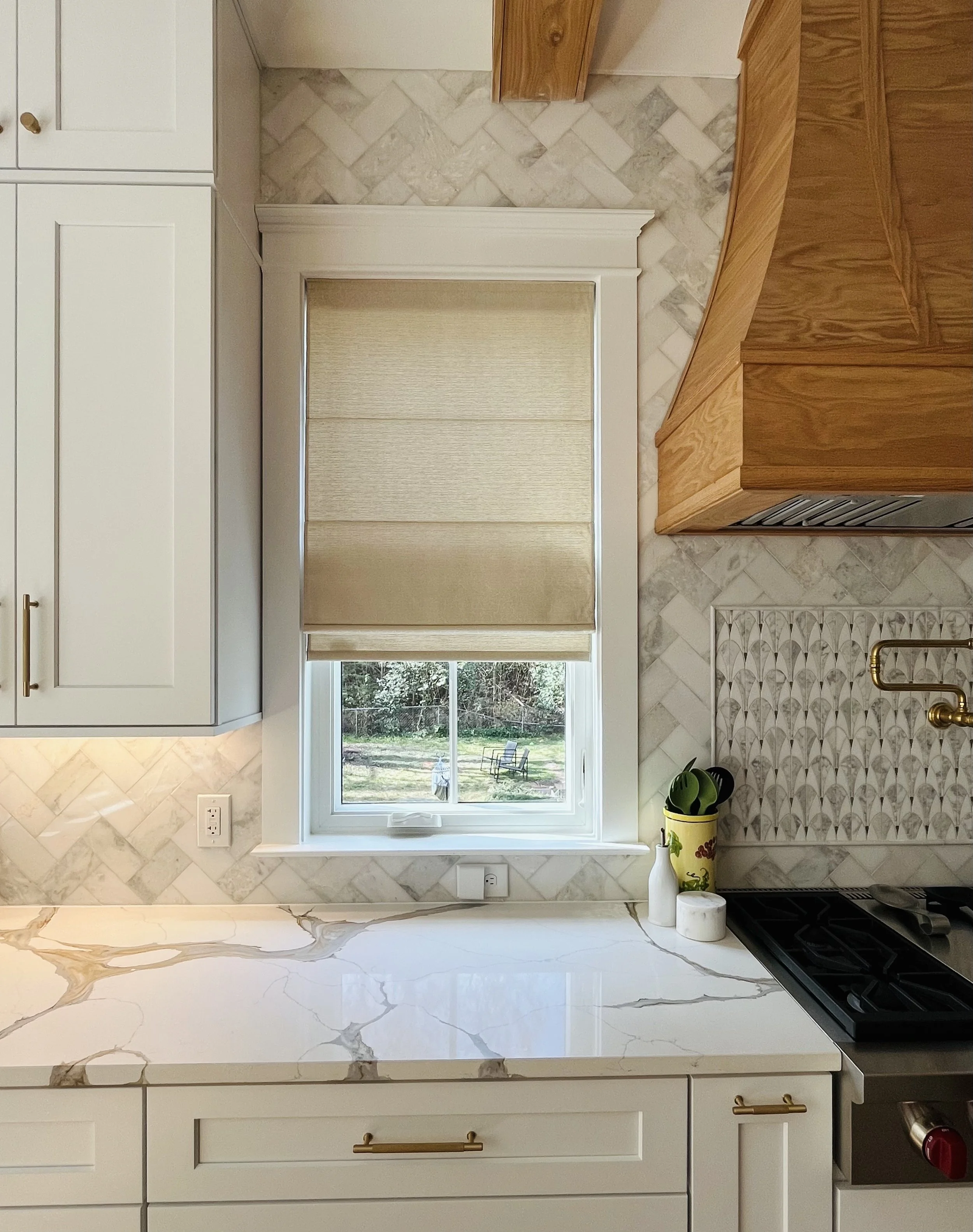 Modern kitchen interior featuring white cabinets, a marble countertop, a window with a beige Roman shade, a gas cooktop with a wooden range hood, and decorative wall tiles.
