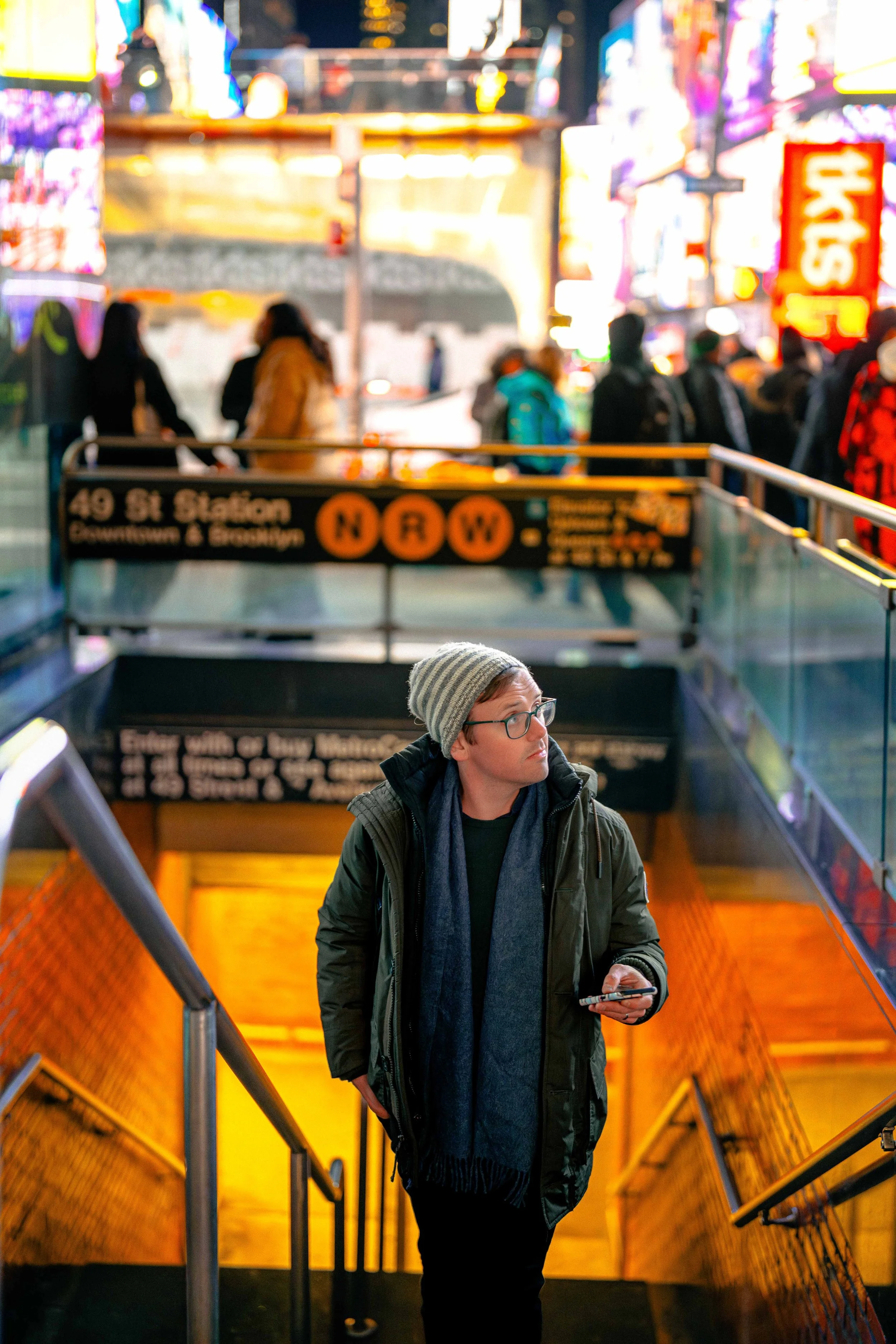 A man with glasses, a striped beanie, and dark winter clothing standing at the entrance to a subway station, holding a smartphone, with a busy, illuminated city street in the background.